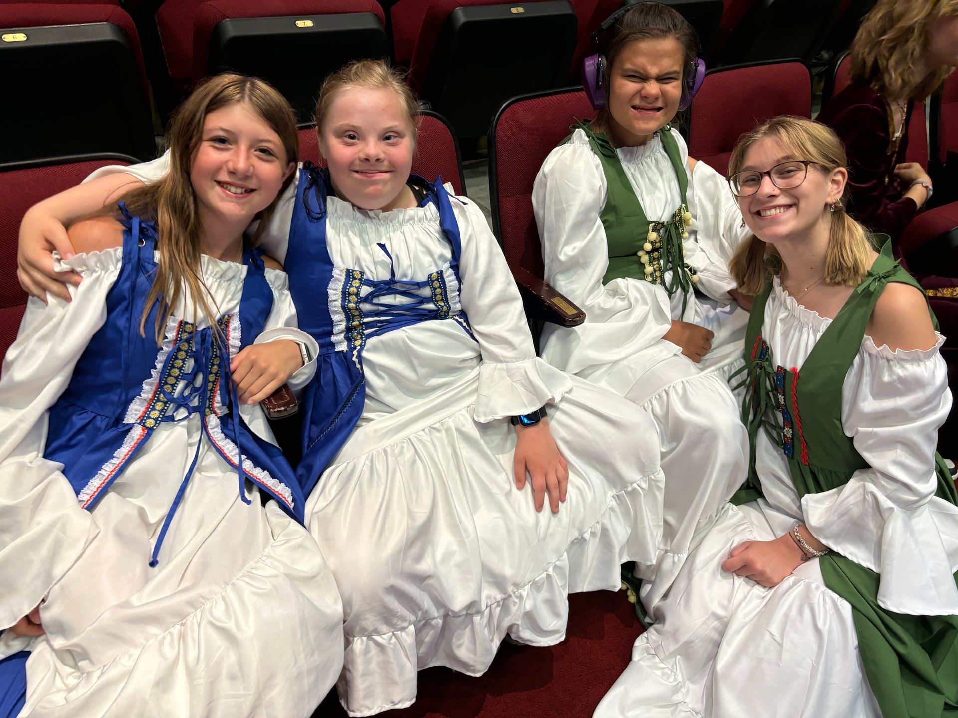 Four young women in Renaissance costumes smiling in theater seats.