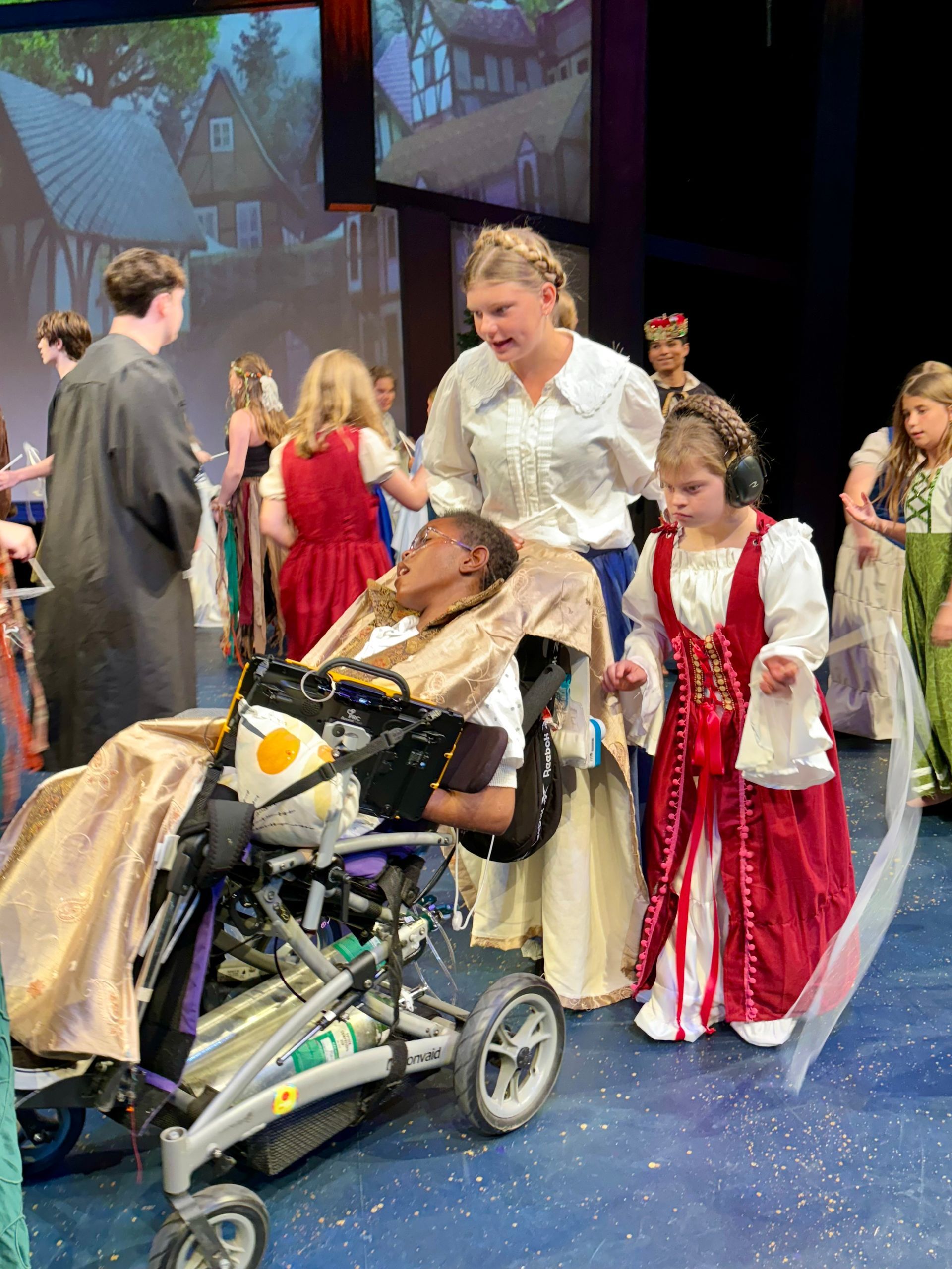 People in period costumes on stage, including one in a wheelchair. Two assist him. Colorful, cheerful setting.