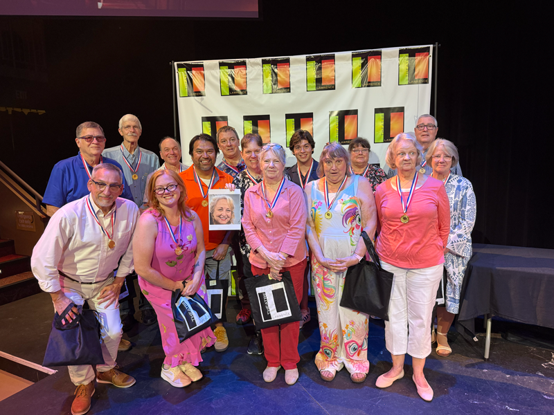 Group of people wearing medals posing for a photo in front of a stage with a colorful banner.