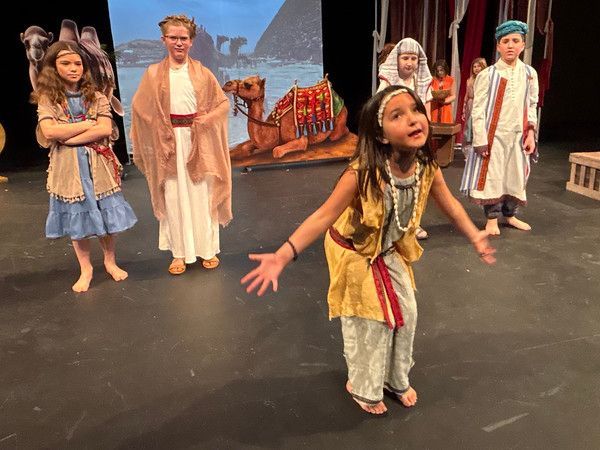 Children in costumes on stage, performing a play with a desert backdrop. One child gestures.