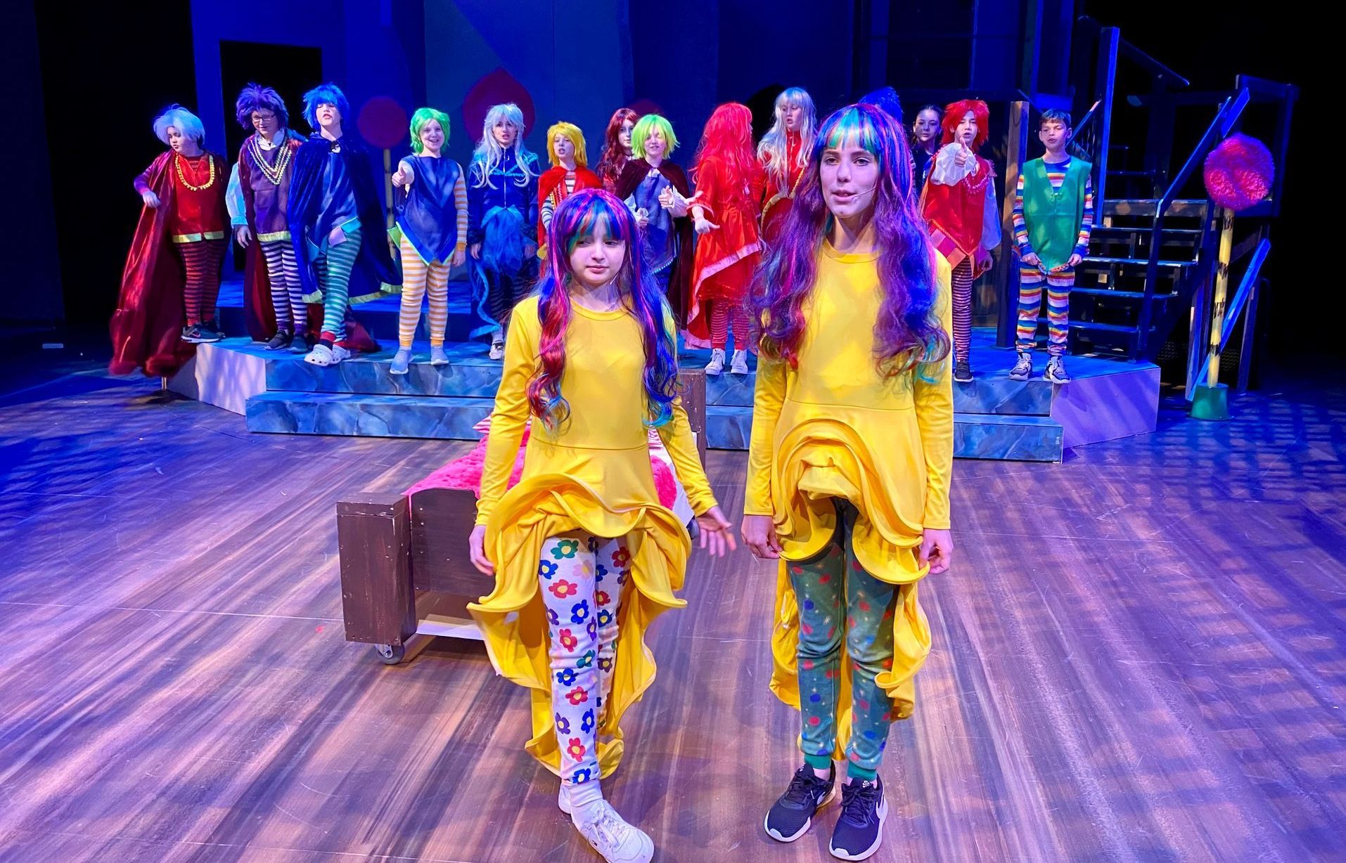Youth production, Two girls in yellow flower costumes on stage, surrounded by colorful costumed performers.