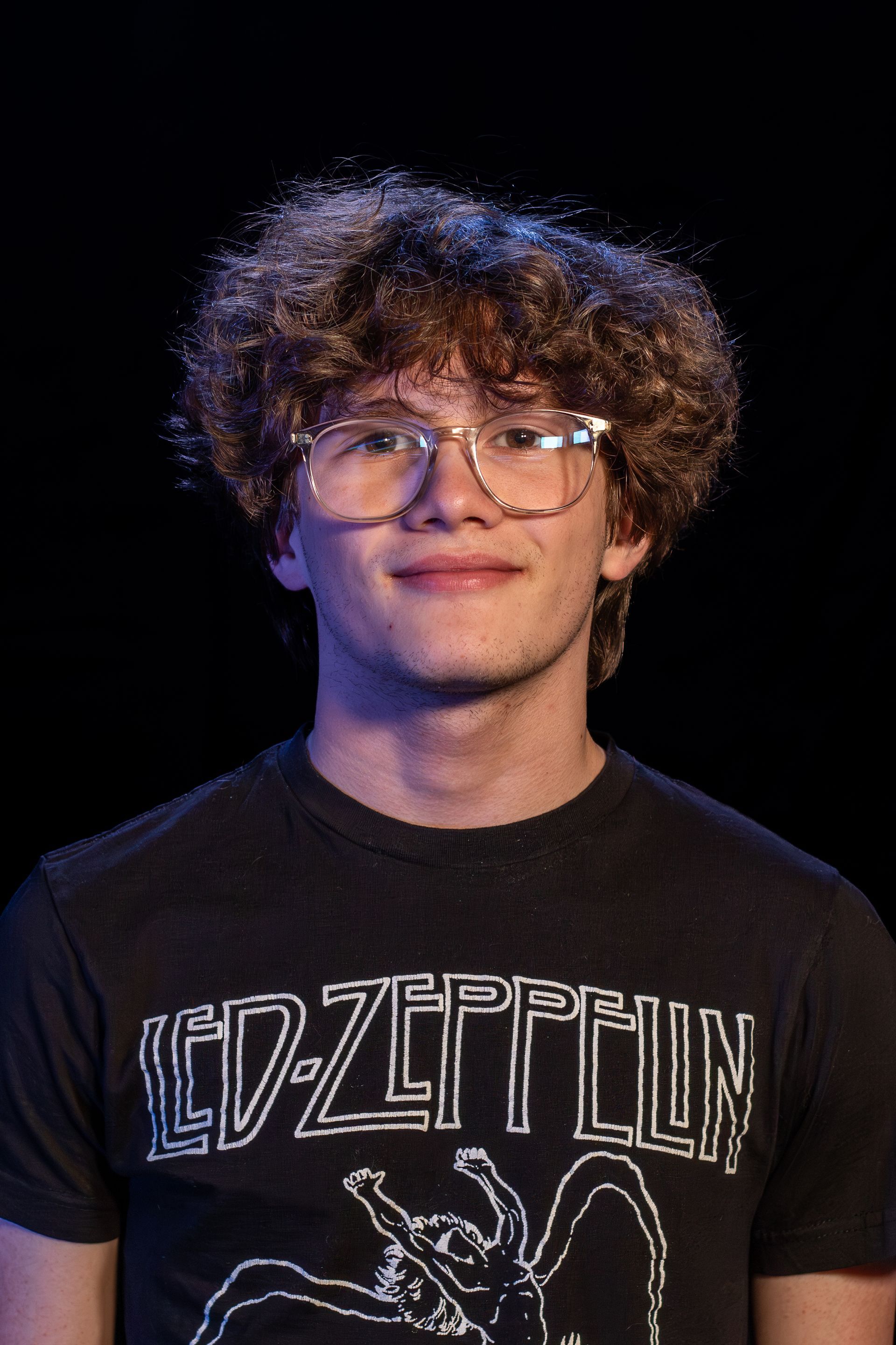 Man with curly hair and glasses wearing a Led Zeppelin t-shirt, smiling, against a dark backdrop.