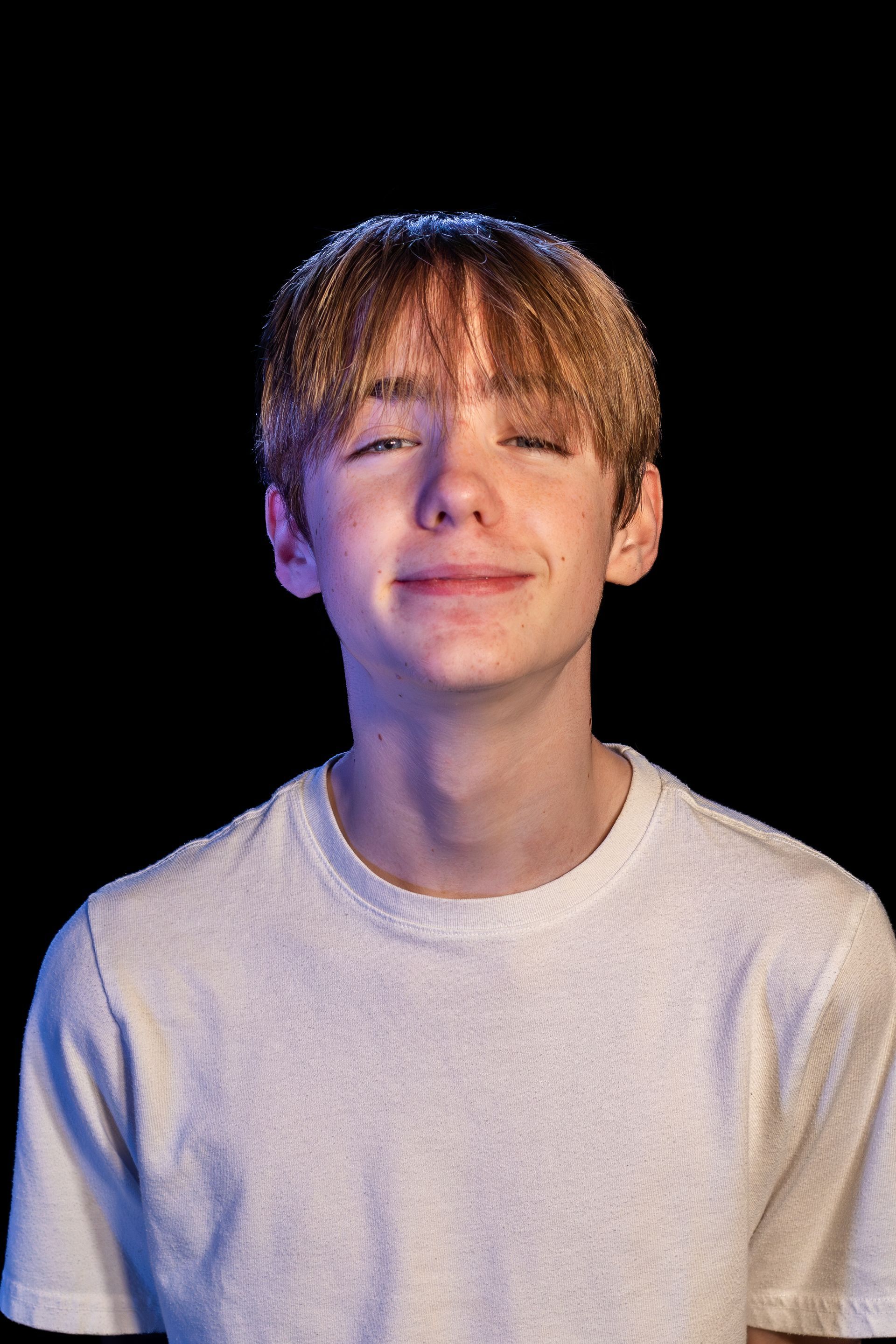 Young person with light brown hair, smiling, wearing a white shirt against a black background.