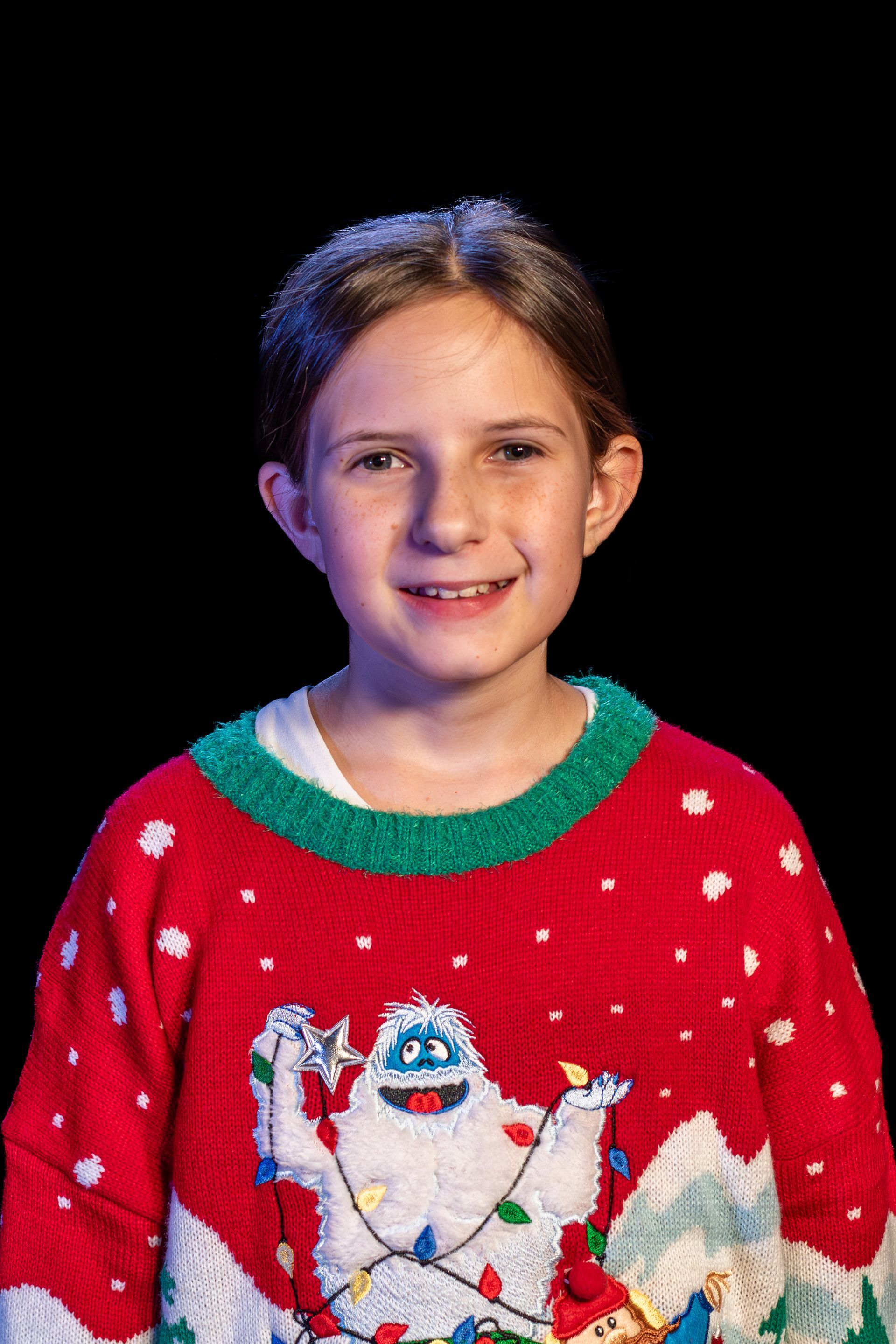 Girl smiling, wearing a red Christmas sweater with a Bumble character on it, black background.