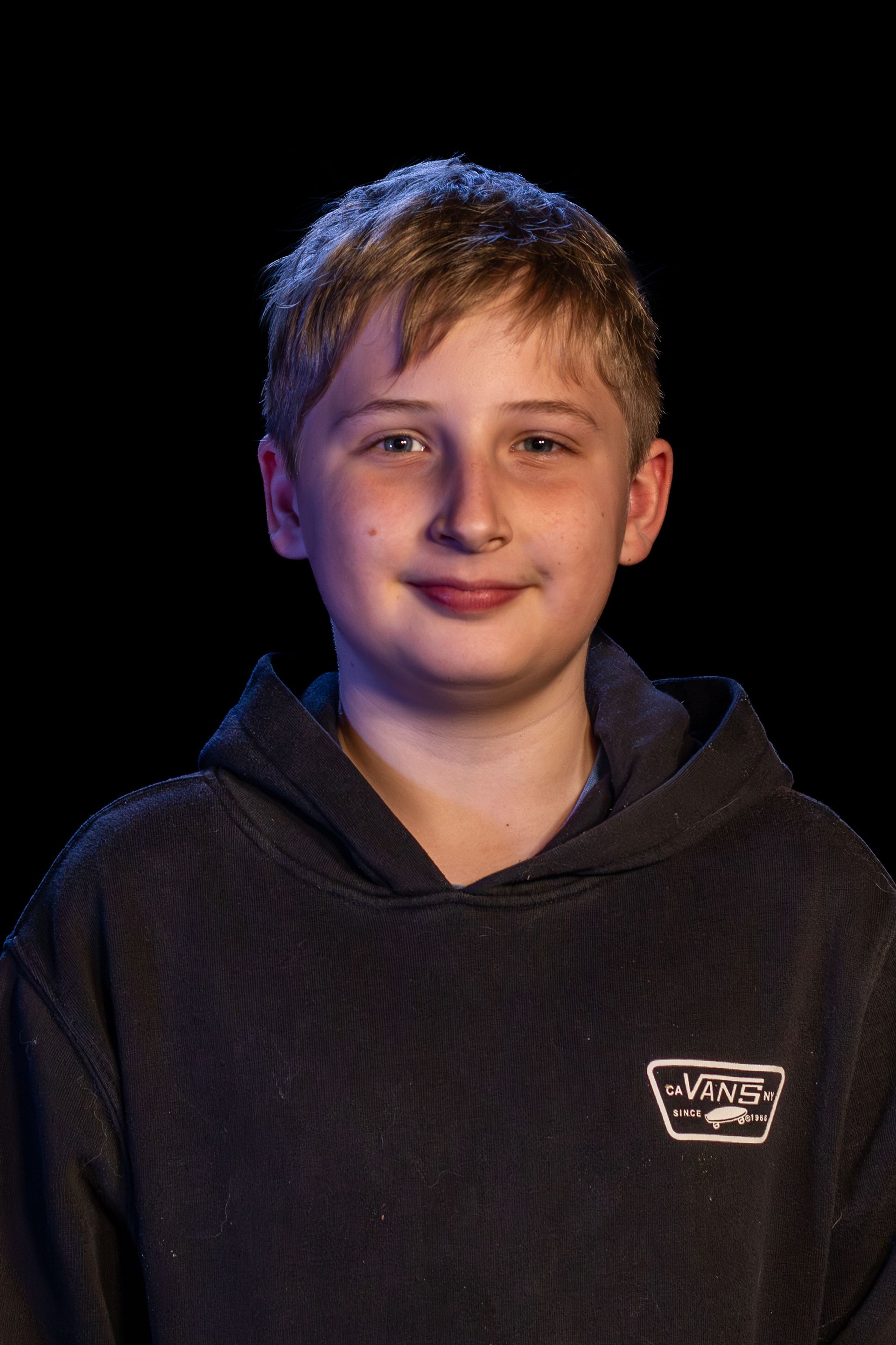 A young person in a dark hoodie smiles at the camera against a black backdrop.