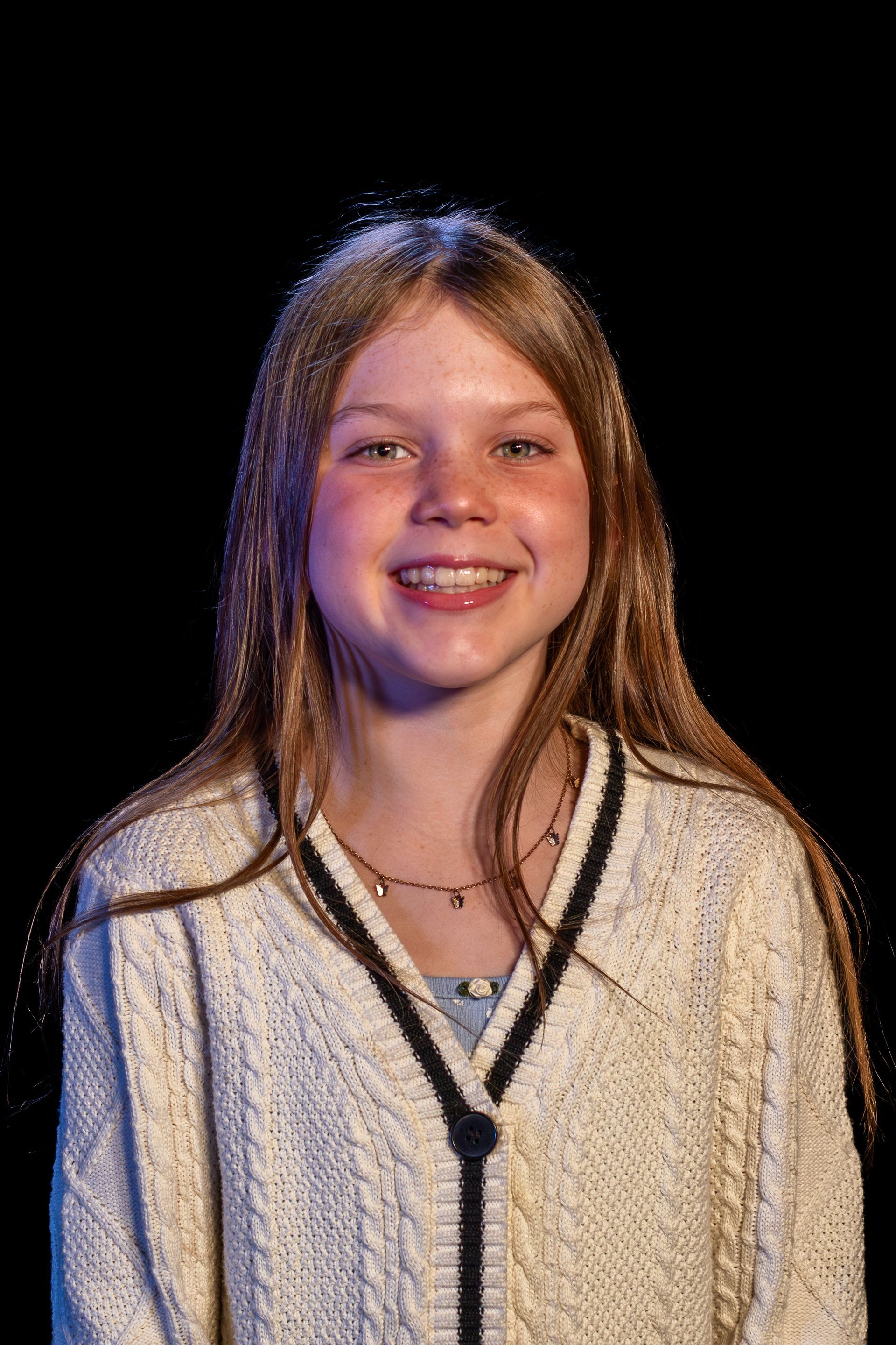 Smiling person with long blonde hair wearing a cream-colored cardigan with a dark trim. Dark background.