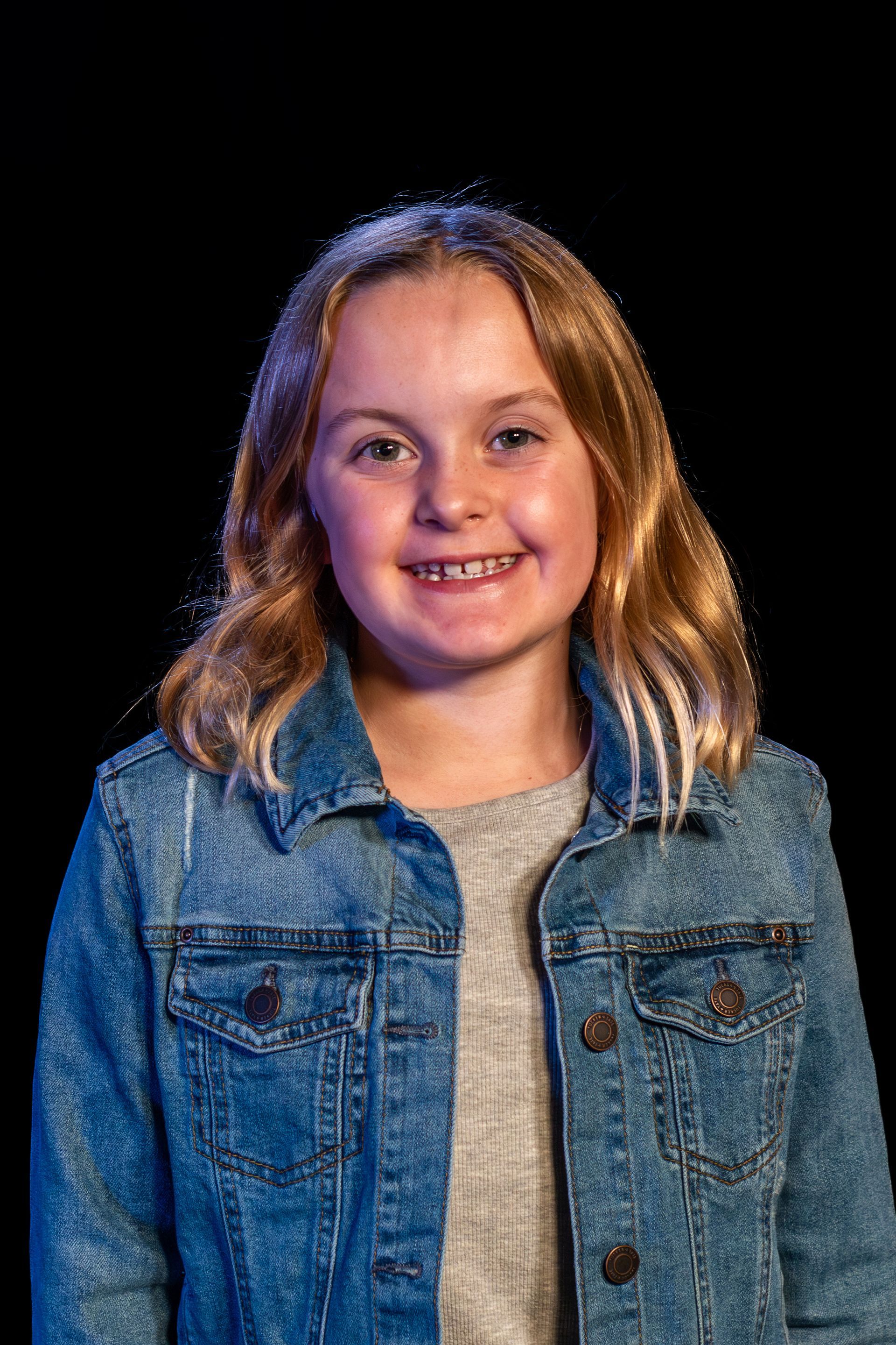 Girl with braces smiles, wearing a denim jacket over a gray shirt. Black background.