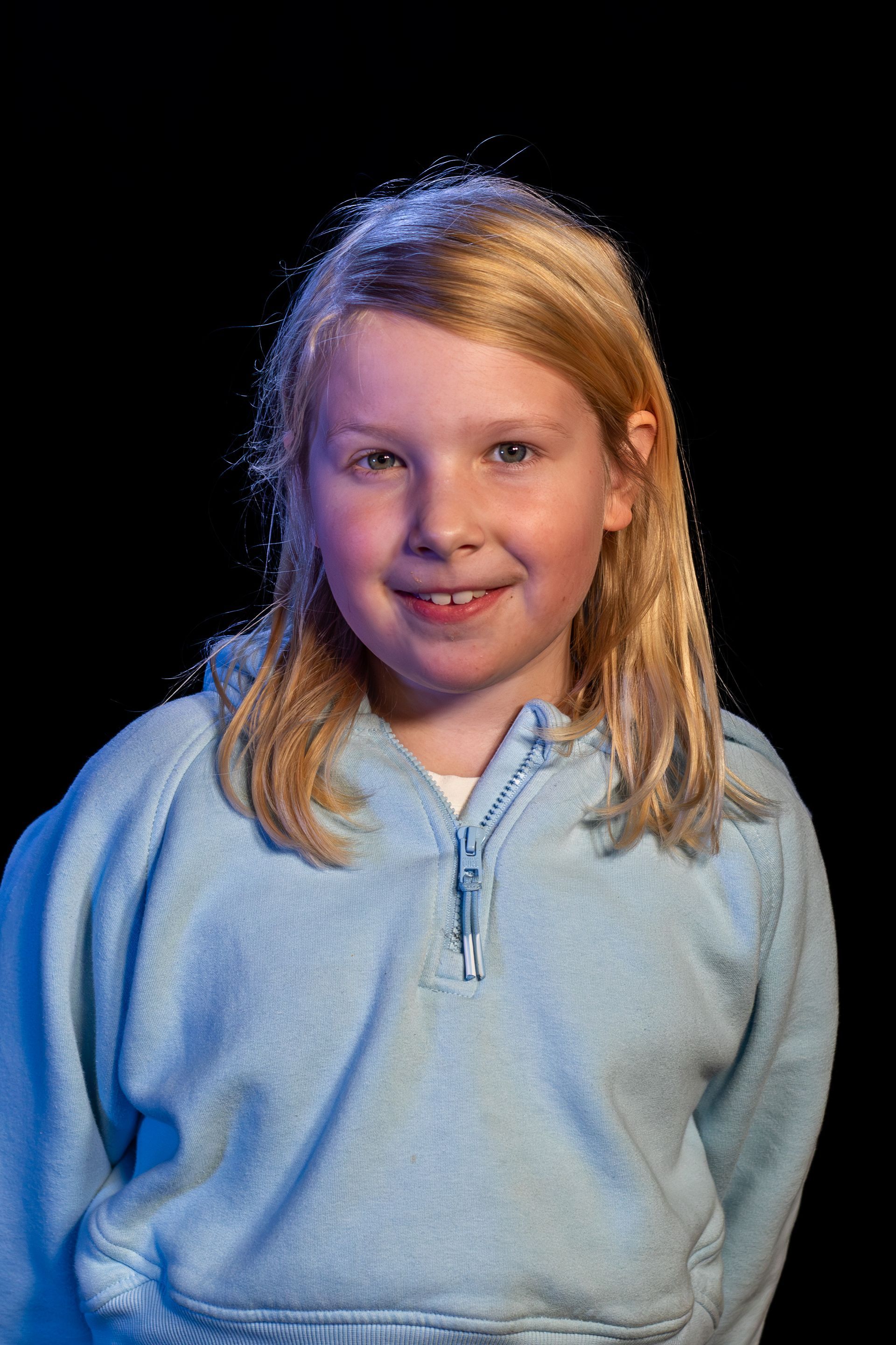 Blonde-haired child in a blue hoodie smiles at the camera against a black background.