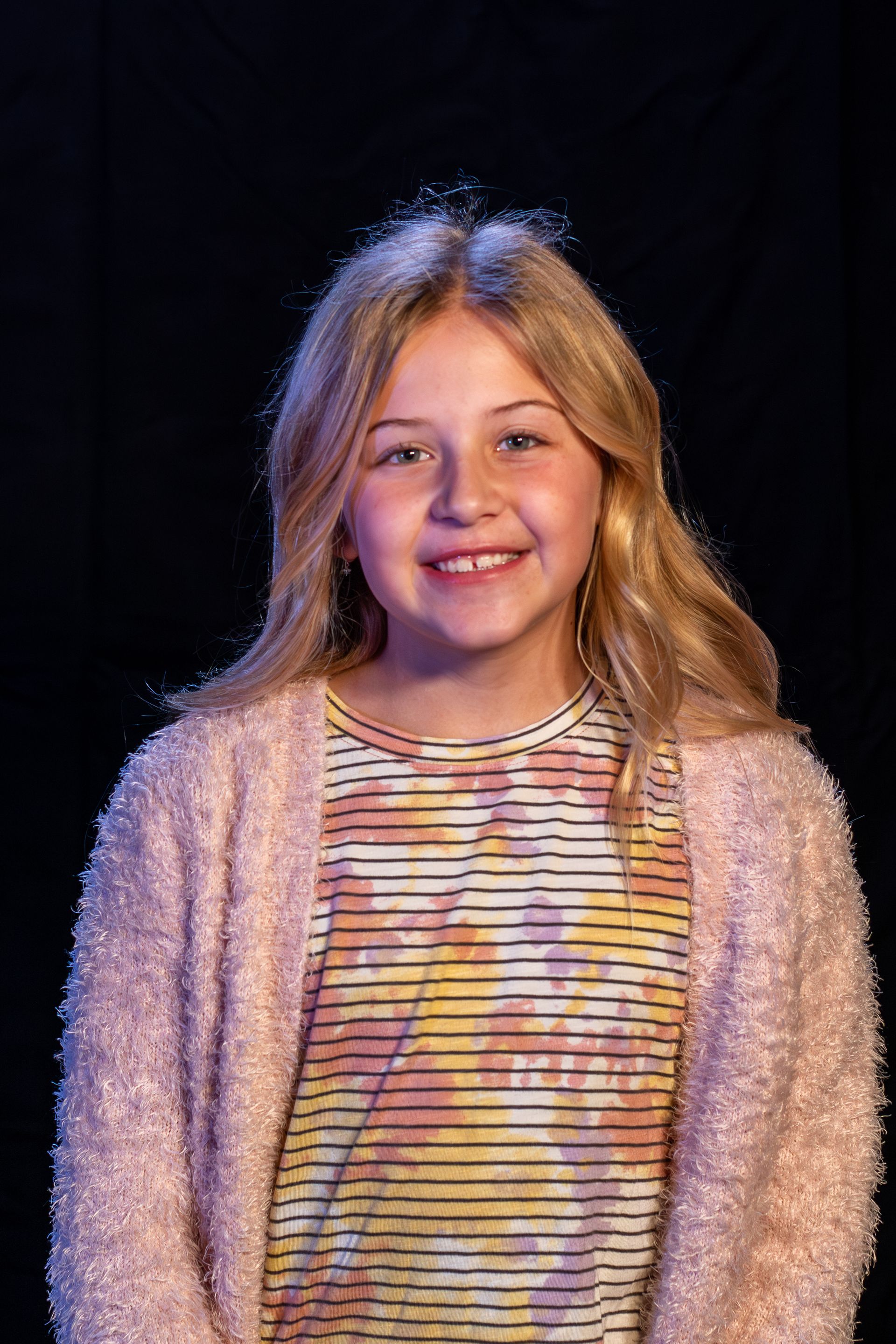 Young person smiles, wearing a striped shirt and pink cardigan, against a dark background.
