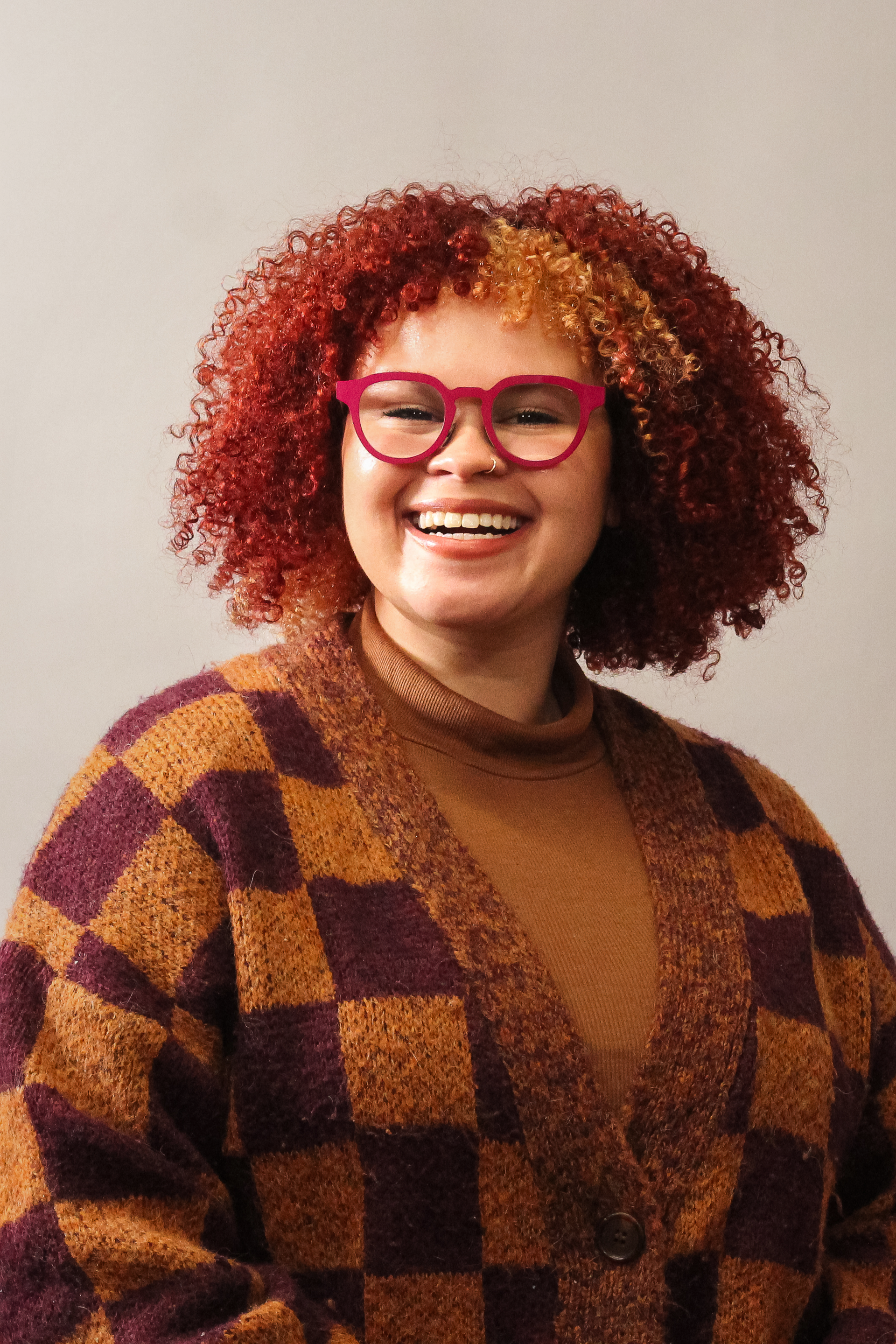 Woman with curly red hair and pink glasses, wearing a checkered cardigan, smiling.