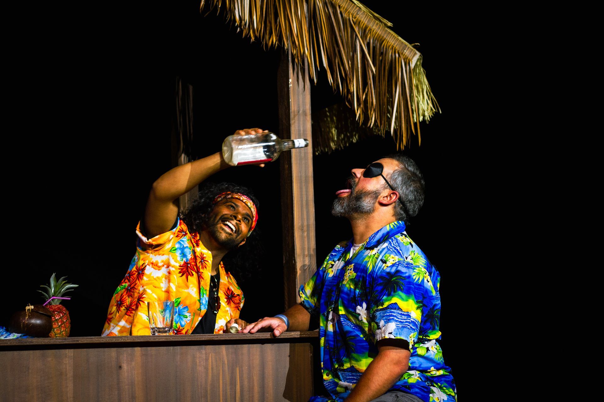 Two people in Hawaiian shirts at a bar. One pours a bottle into the other's mouth. Dark background.