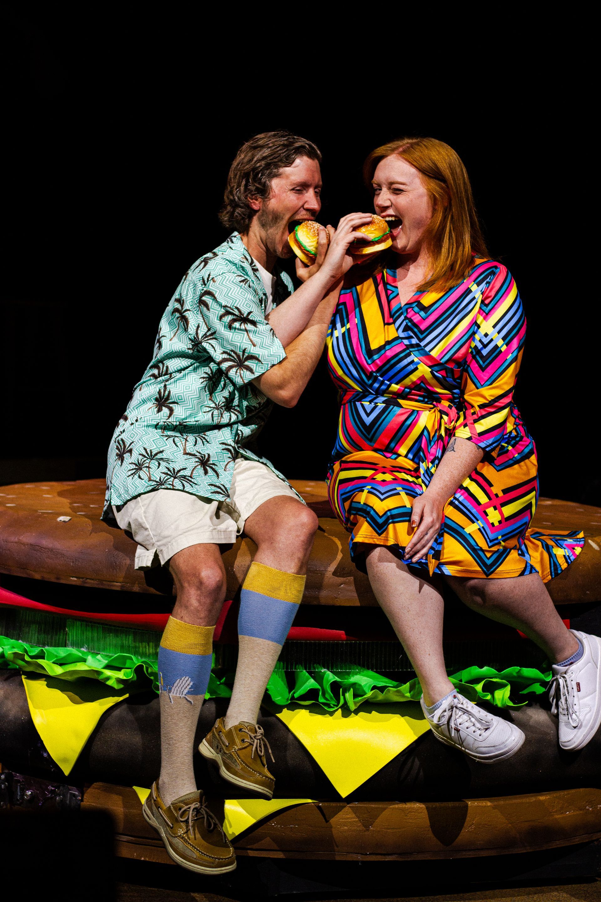 Two people eating giant burger outdoors; man in shorts/Hawaiian shirt; woman in colorful dress.