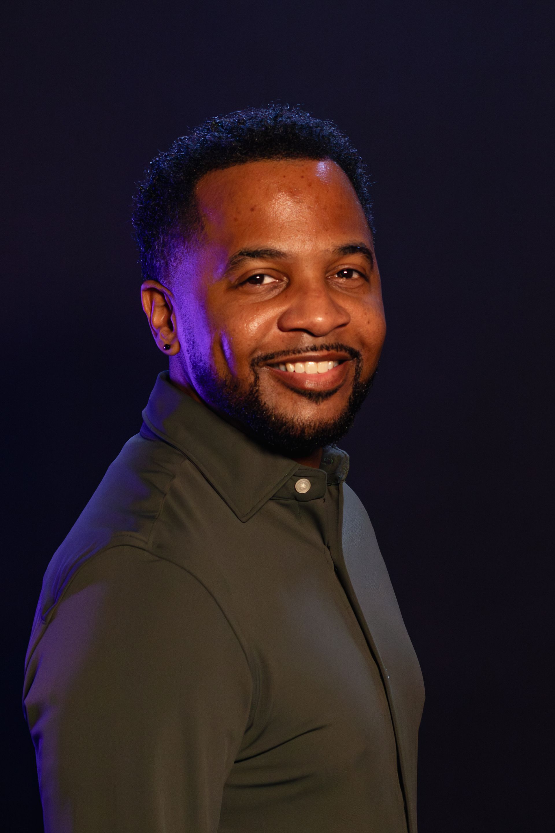 Man with a beard smiling, wearing a dark green shirt, on a dark blue background.