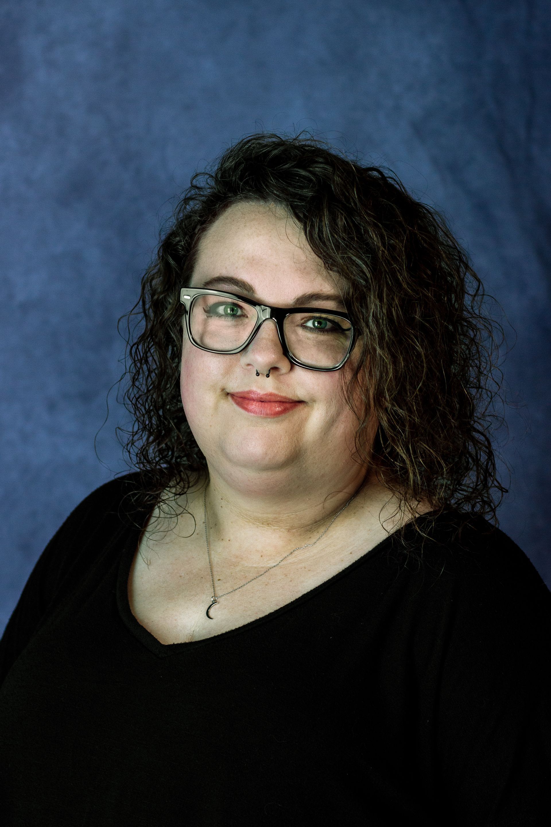 Woman with glasses smiles, wearing black shirt, against blue background.