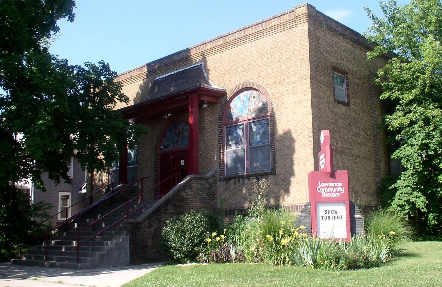 Brick building with red trim, steps, and sign, possibly a church, set in a grassy area.
