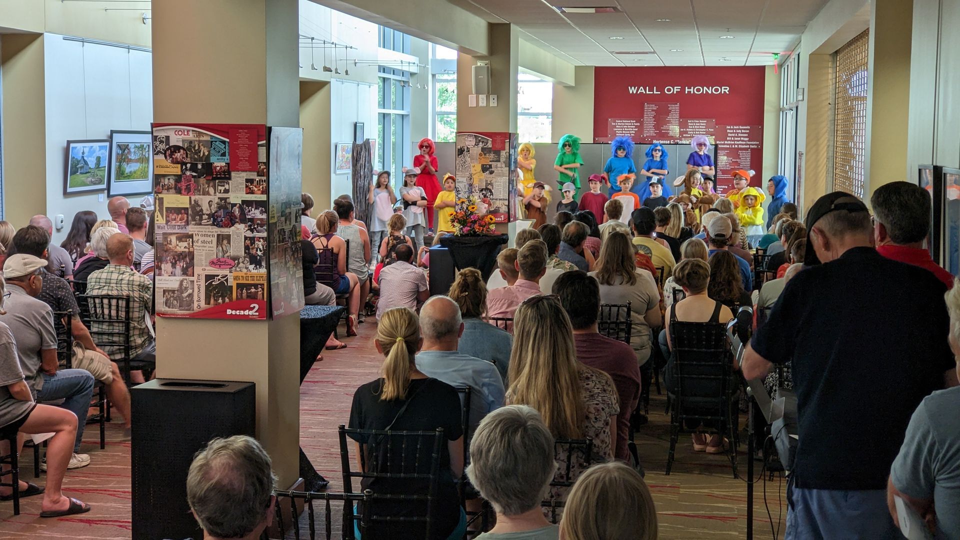 Audience watches costumed characters on a stage in a crowded hall; framed photos and banners on the walls.