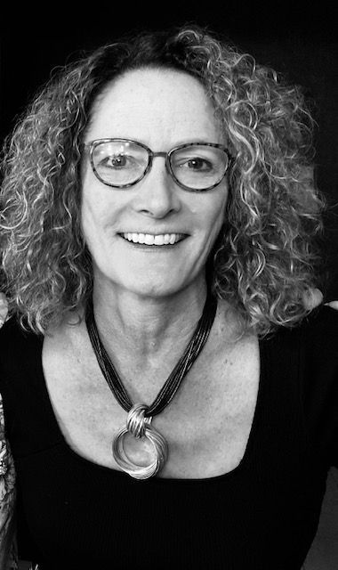 Woman with curly hair, glasses, smiling, wearing a necklace.