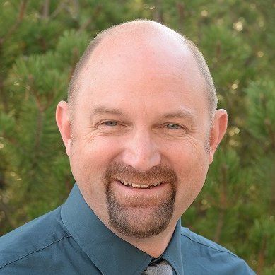 Smiling bald man with a goatee wearing a blue shirt, outdoors with green foliage behind him