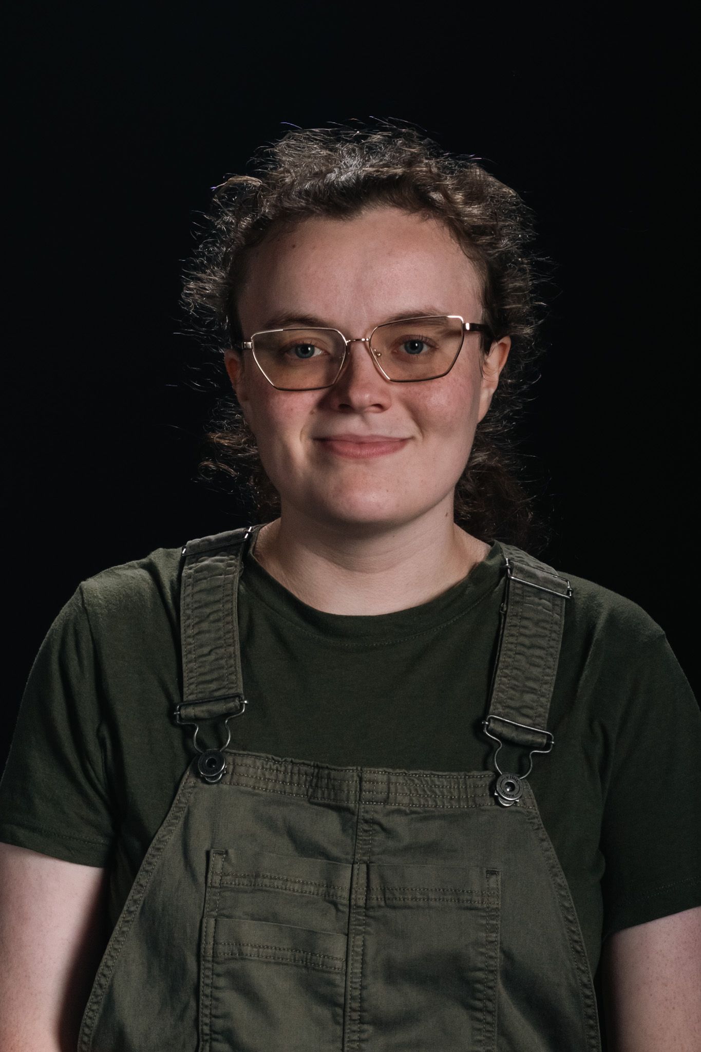 Person wearing glasses and green overalls, facing forward against a dark background.