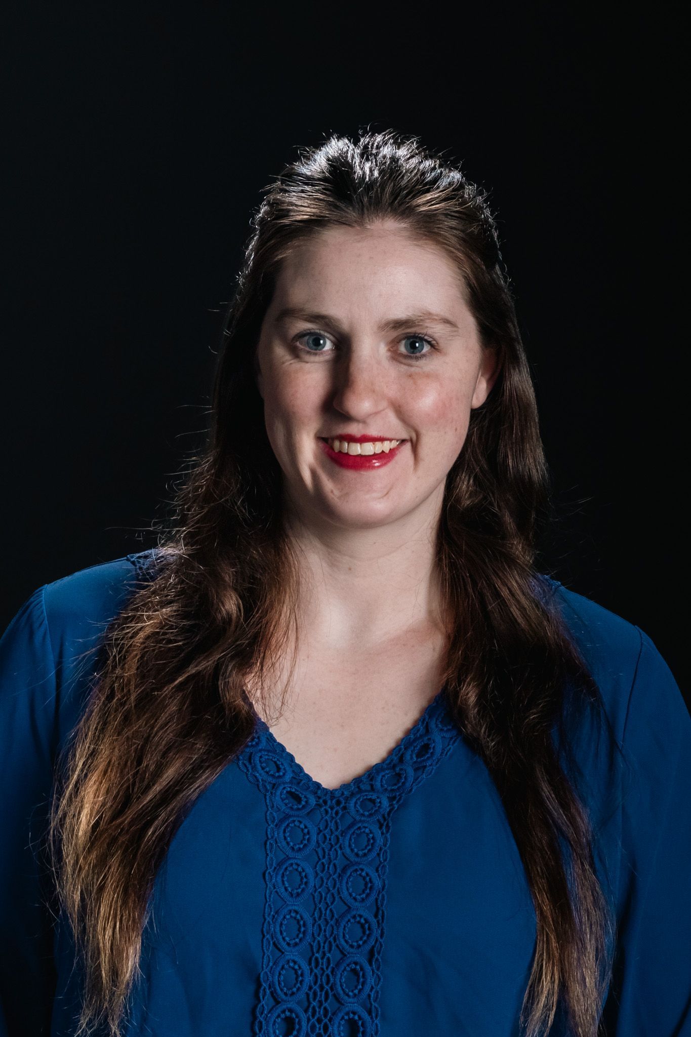 Woman with long dark hair wearing a blue top, smiling against a black background