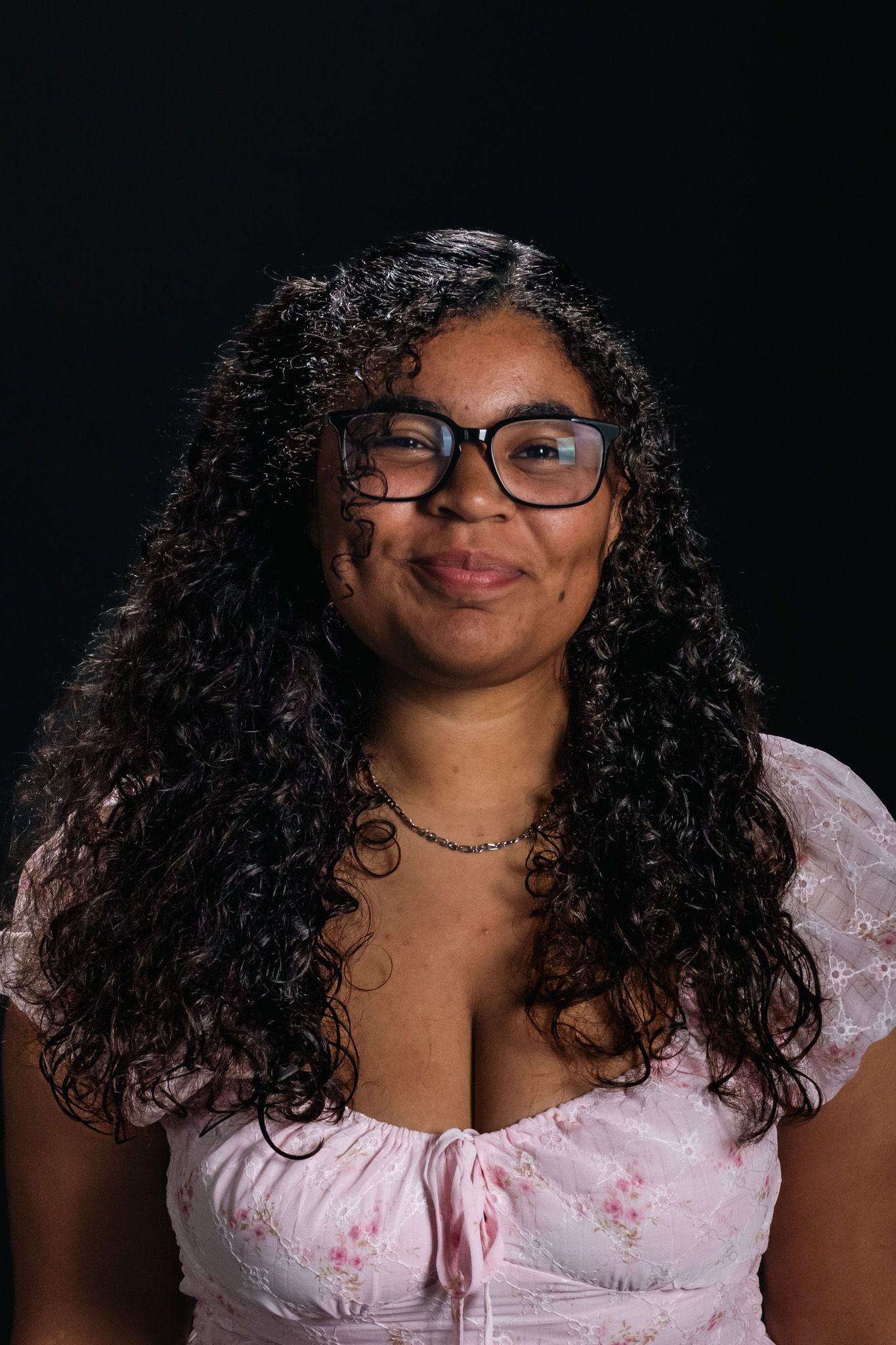 Portrait of a smiling person with curly hair and glasses, wearing a pink top against a dark background