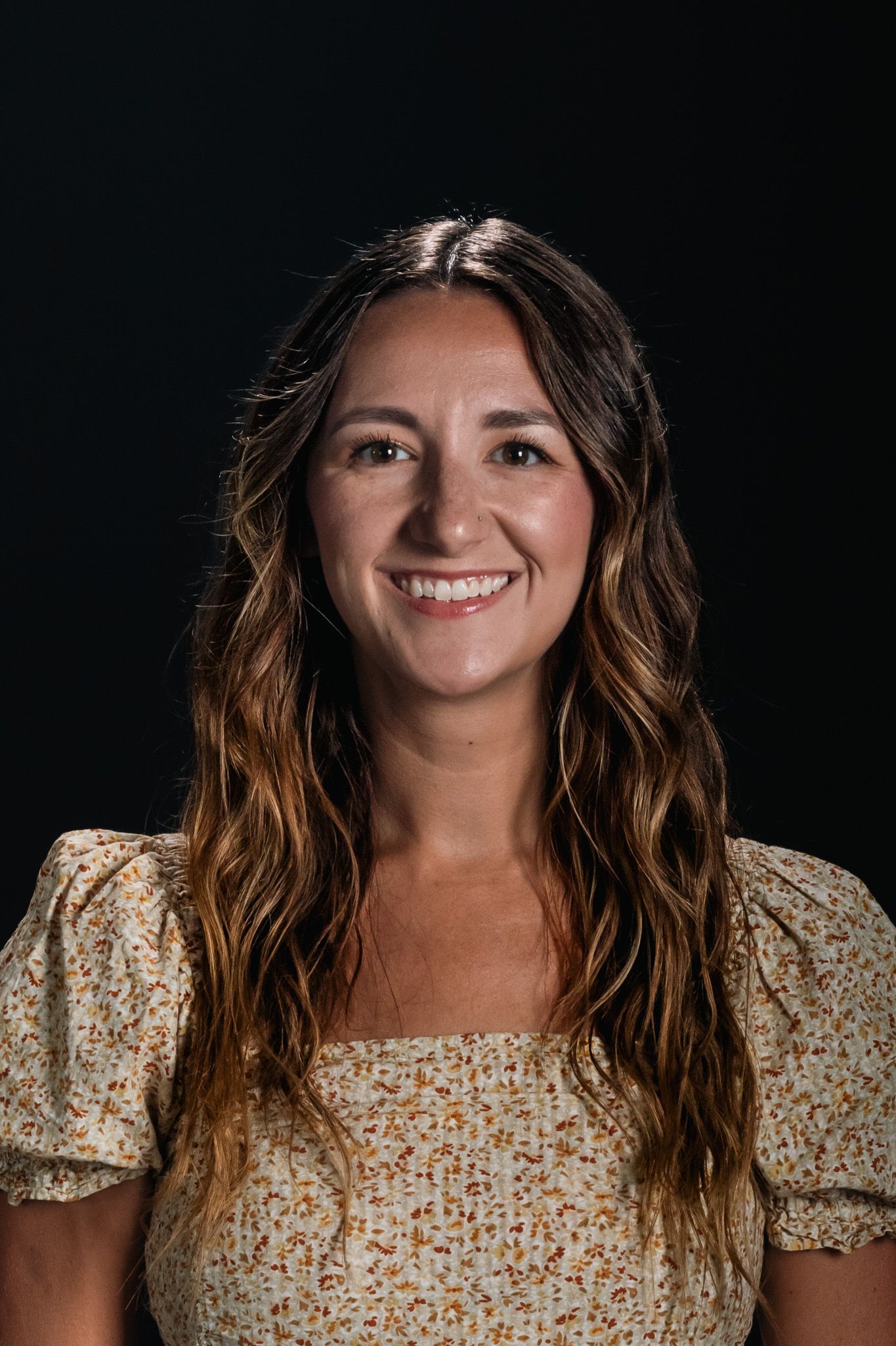 Smiling woman with long wavy hair wearing a patterned blouse against a dark background