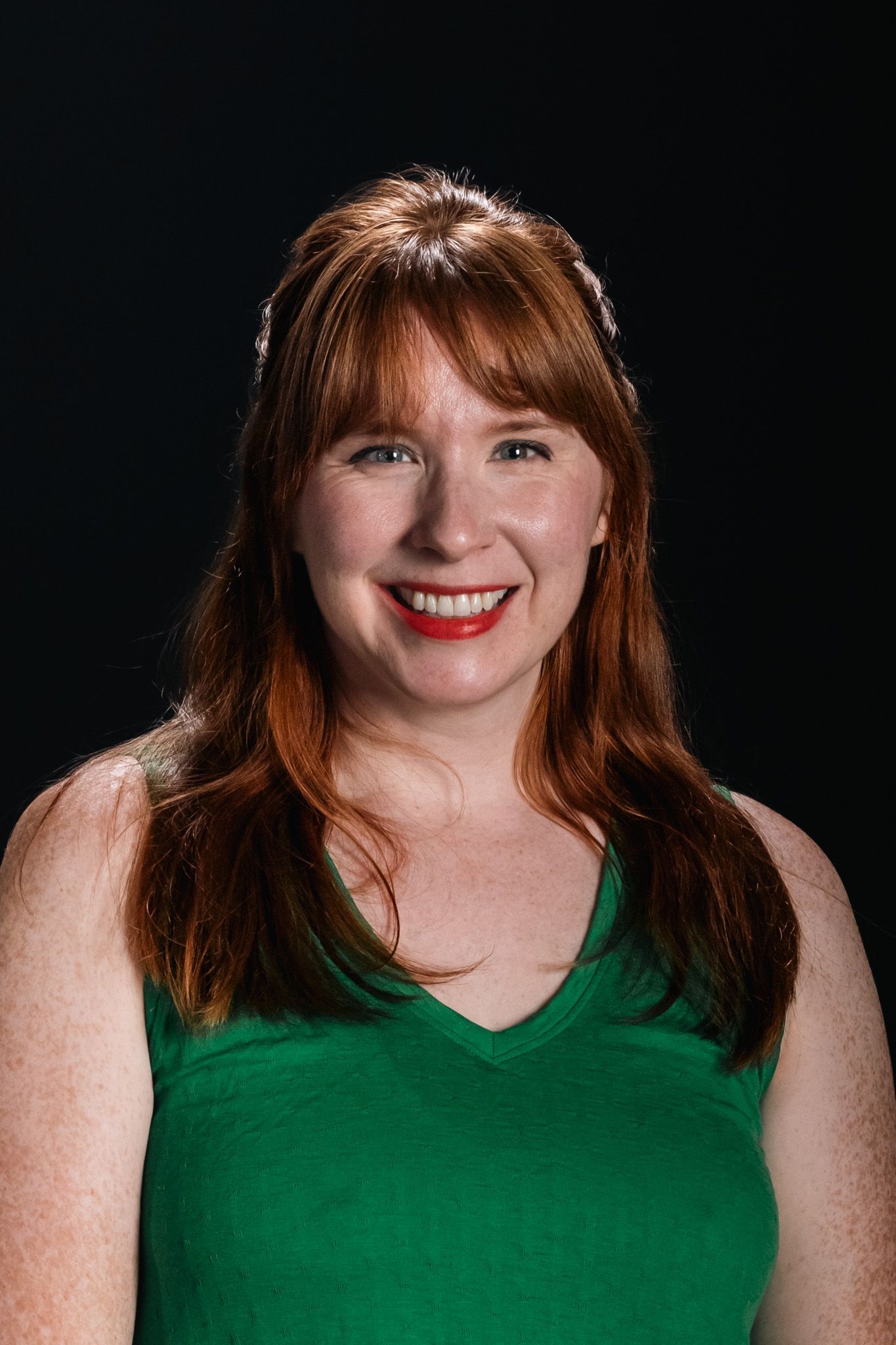 Woman with long brown hair in a green sleeveless top, smiling against a black background