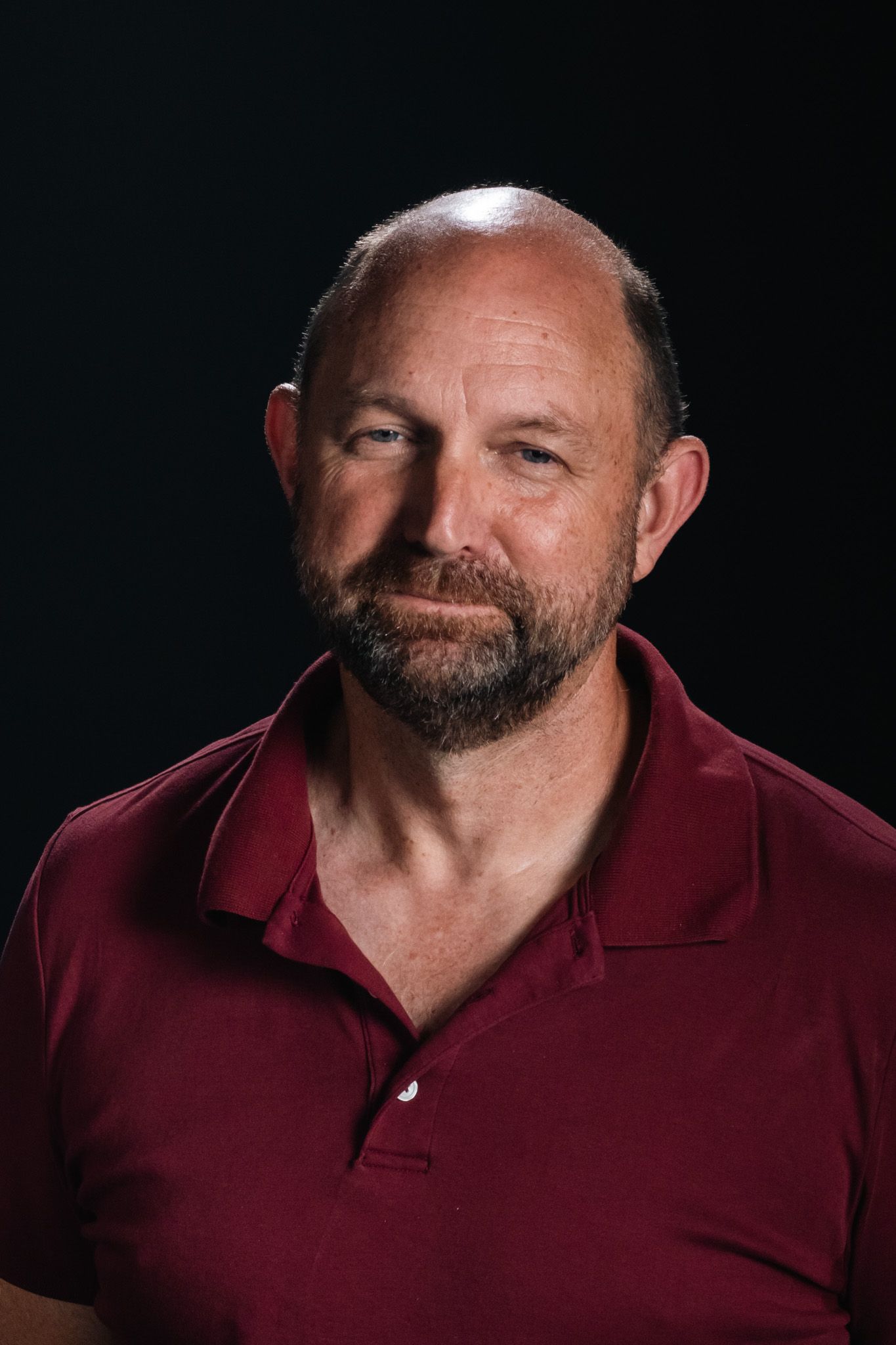 Portrait of a bald man with a beard wearing a maroon polo against a black background