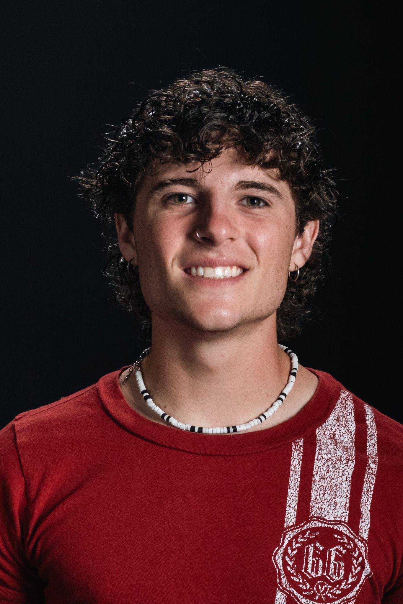 Smiling person in a red shirt with a white graphic, wearing a beaded necklace against a dark background.
