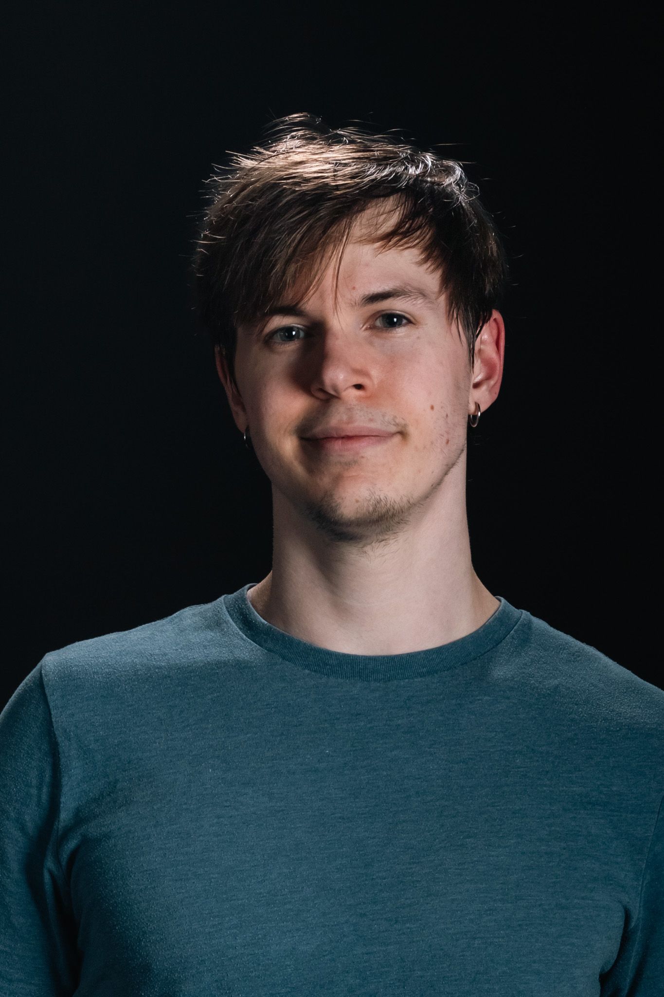 Portrait of a person in a teal shirt against a dark background, looking at the camera with a slight smile