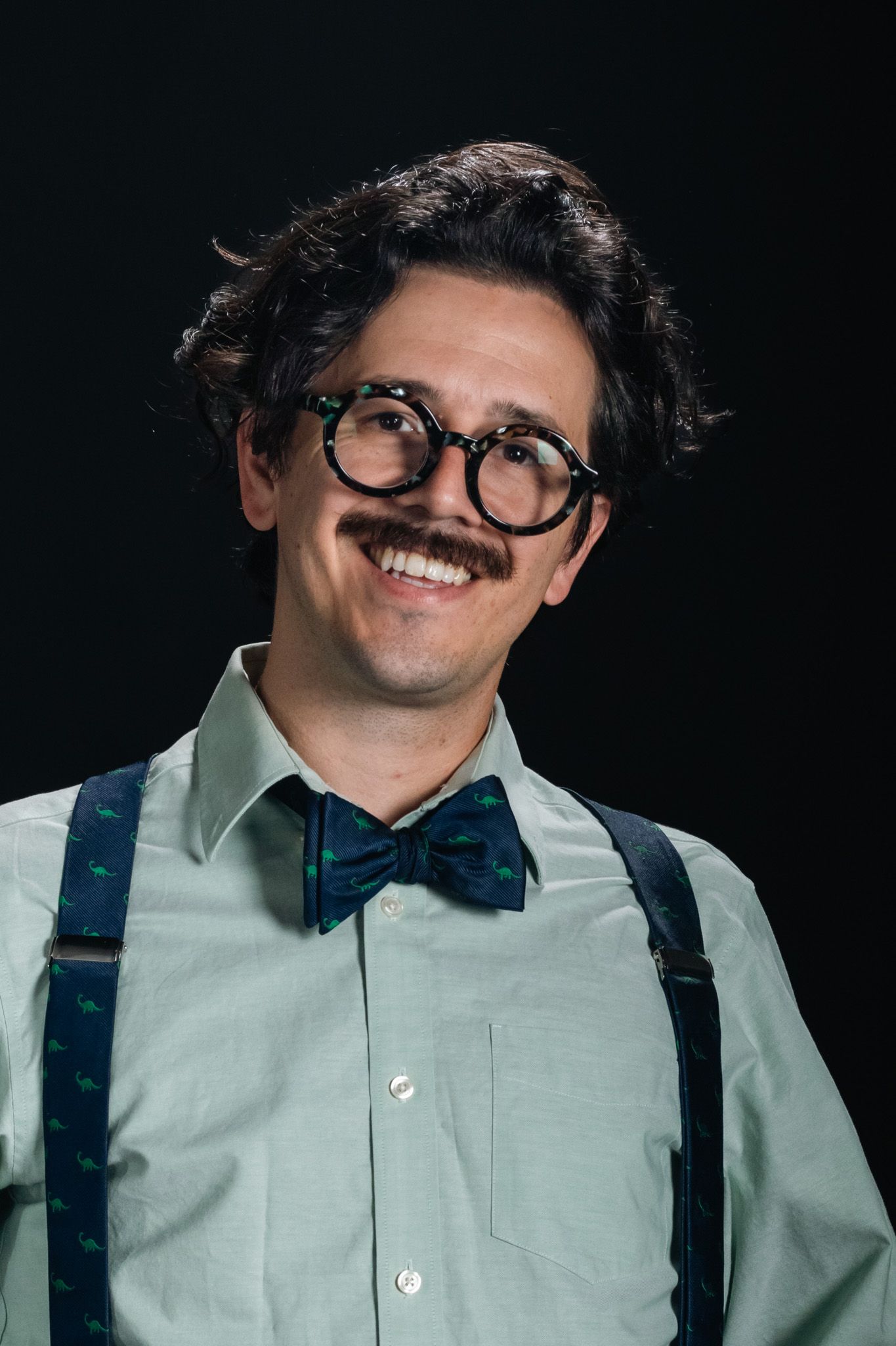 Smiling person with glasses, bow tie, and suspenders against a dark background