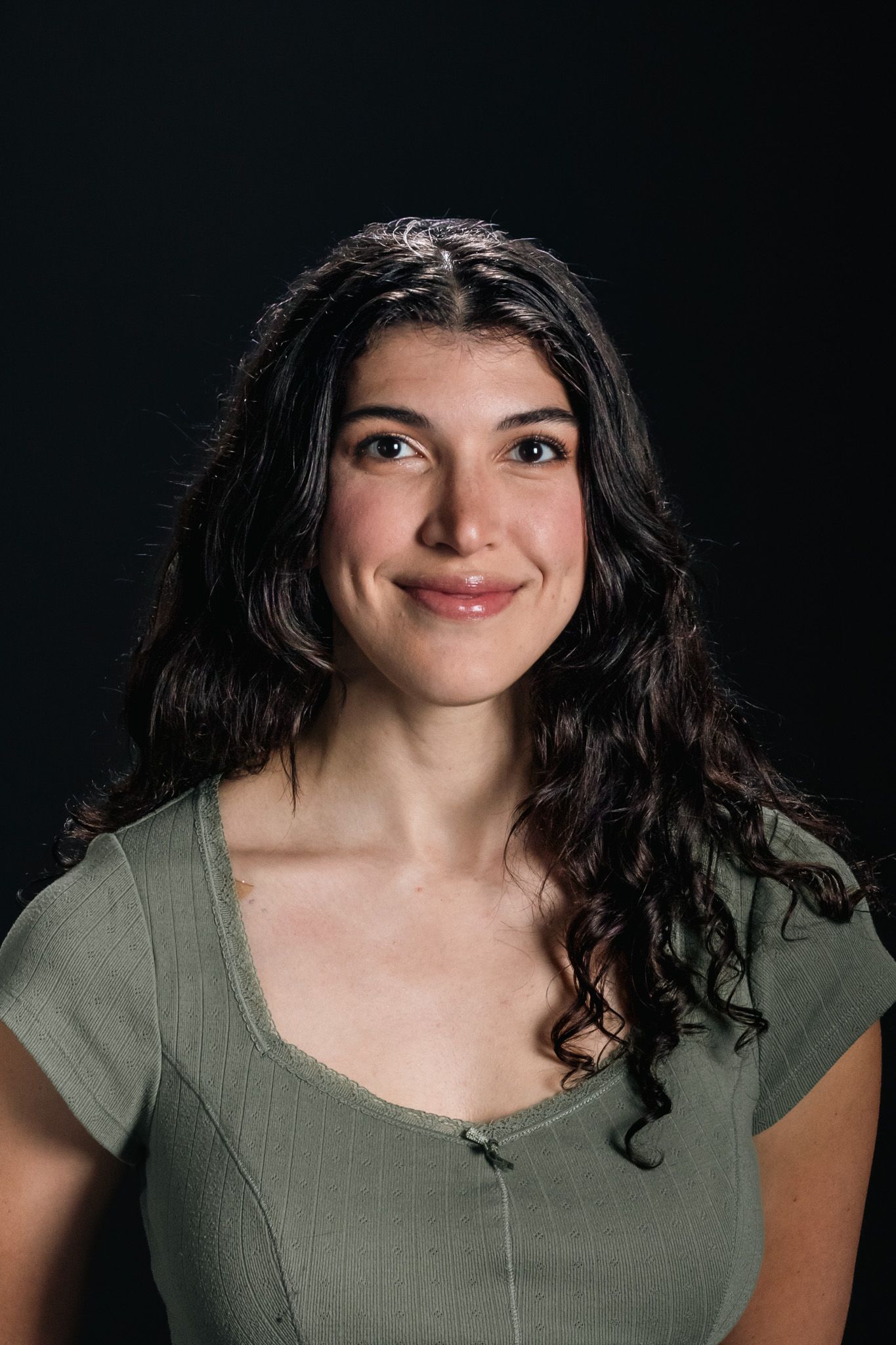 Woman with long dark hair in a green shirt, smiling against a dark background.