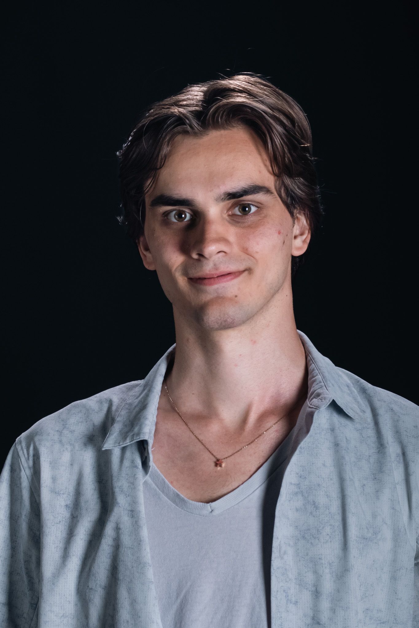 Portrait of a young person with dark hair, wearing a light shirt and necklace against a black background.
