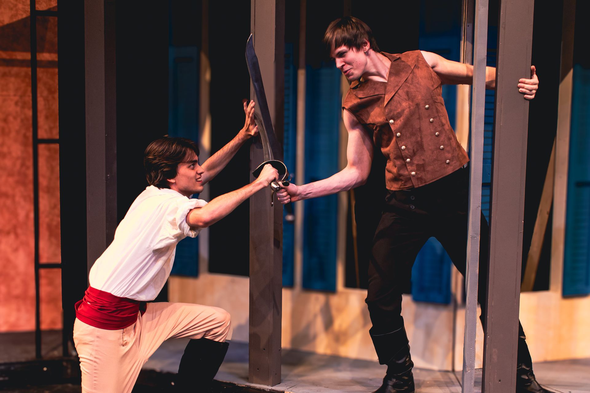 Two actors on stage in costume, reaching toward each other through vertical bars during a dramatic scene