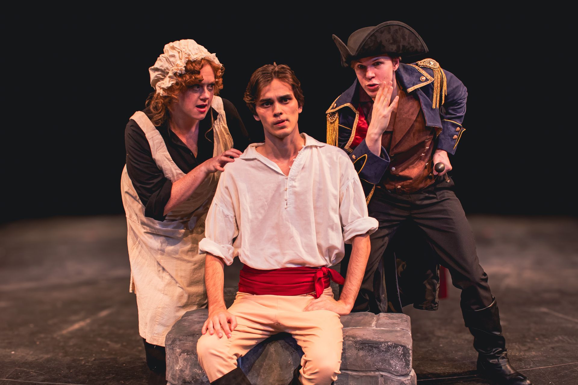 Three people in pirate costumes pose on a dark stage, with one seated on a crate.