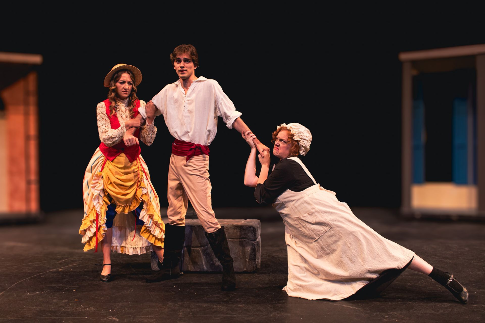 Three actors on a theater stage, one kneeling and reaching toward two standing performers in period costumes.