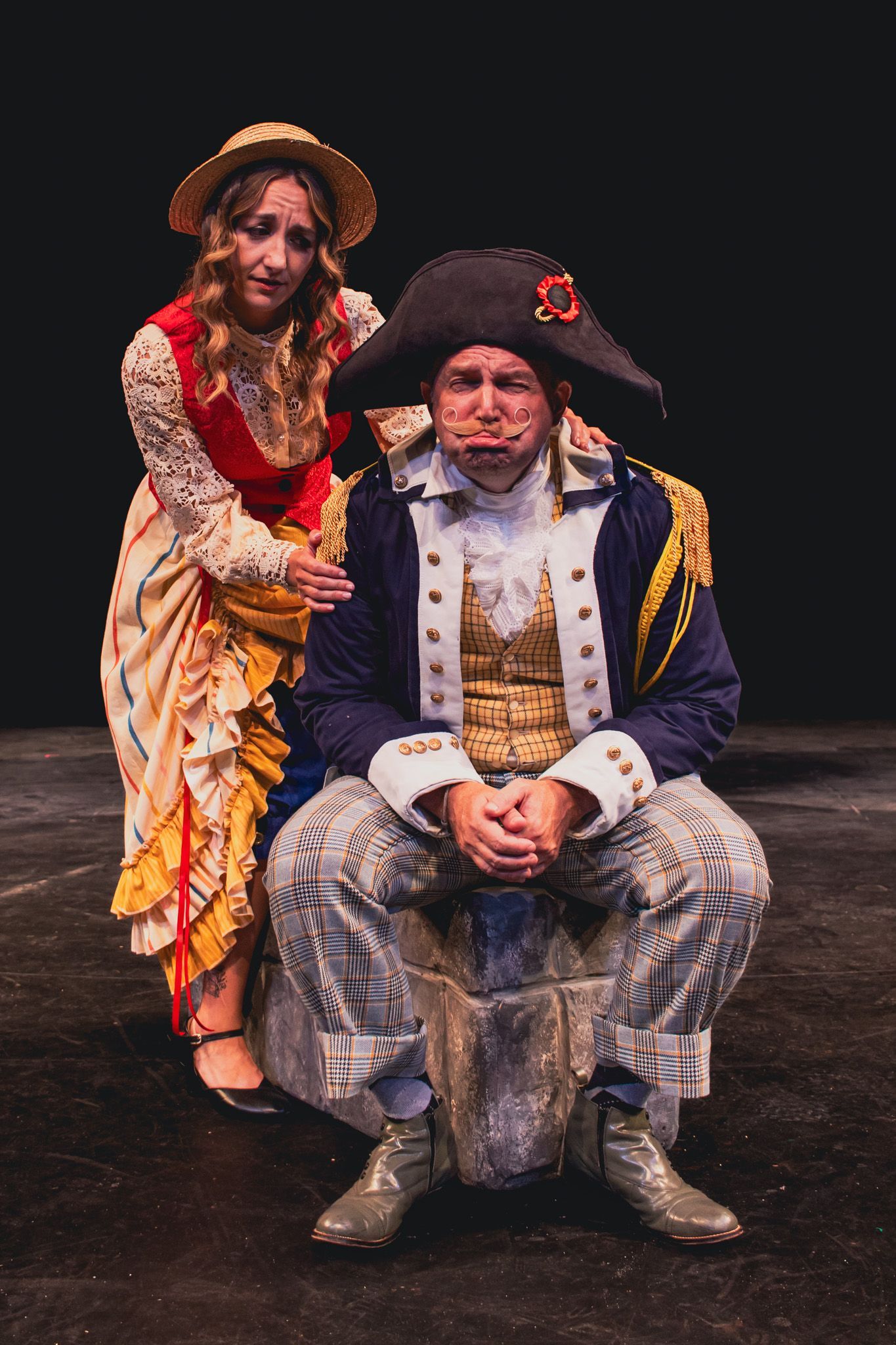 Two actors in ornate 18th-century costumes posing on a dark stage, one seated and one standing behind him