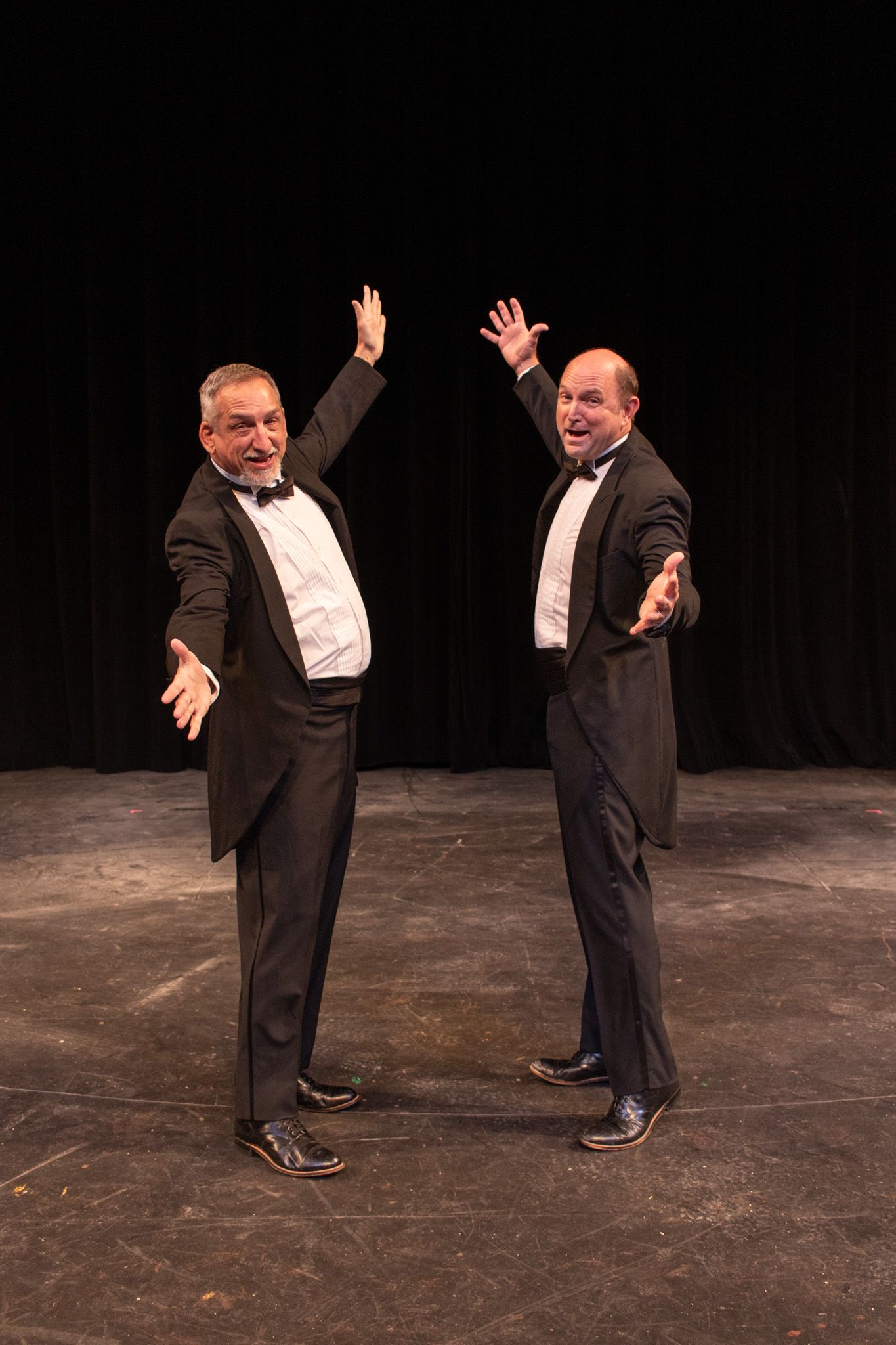 Two men in black suits pose on a dark stage with arms raised in a playful dance pose