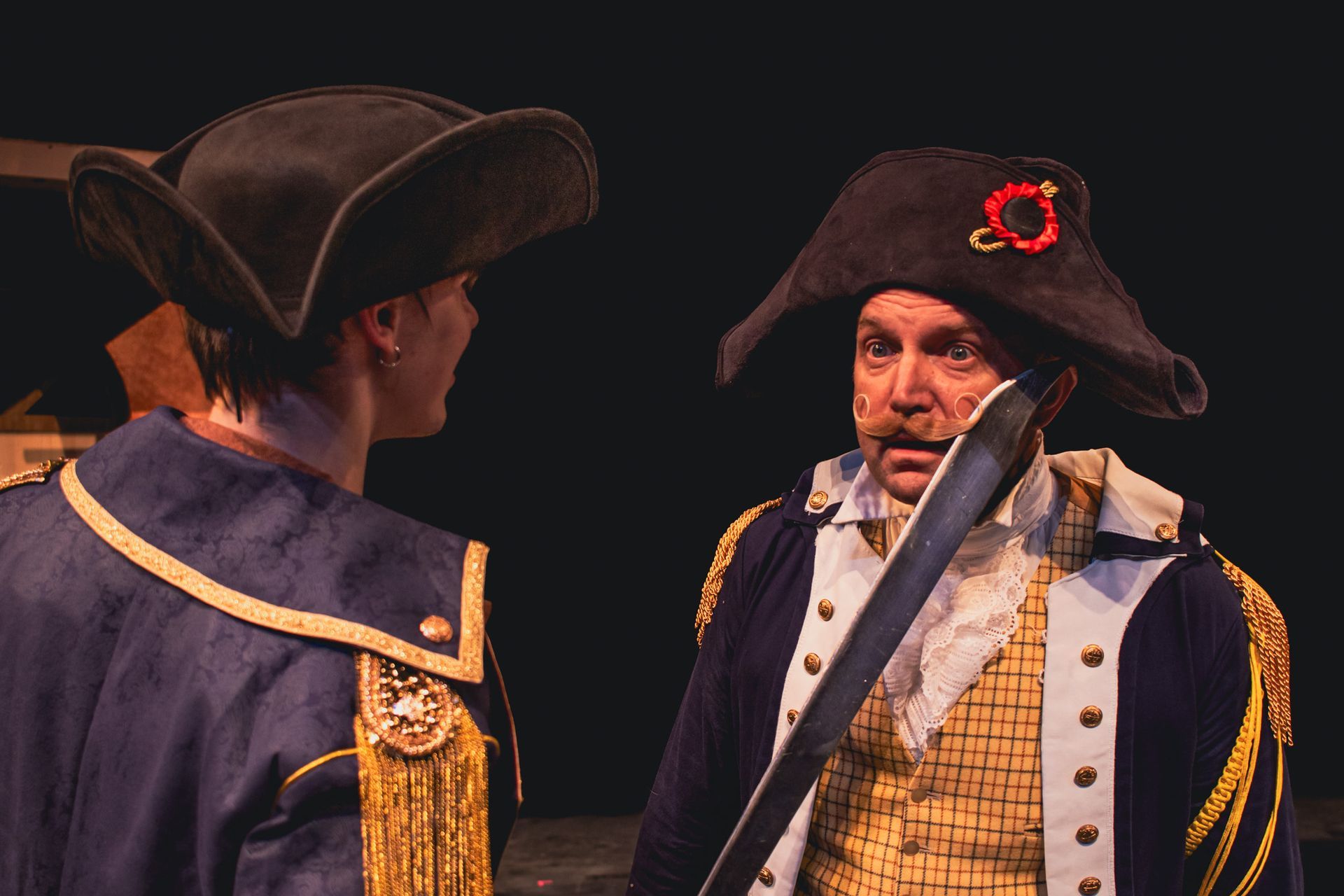 Two men in 18th-century military costumes facing each other on stage, one holding a sword.