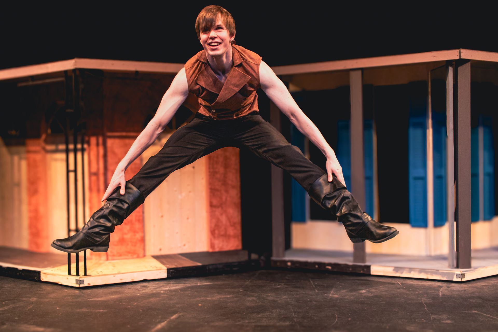Person mid-jump with legs spread on a stage set, wearing dark clothes and smiling.
