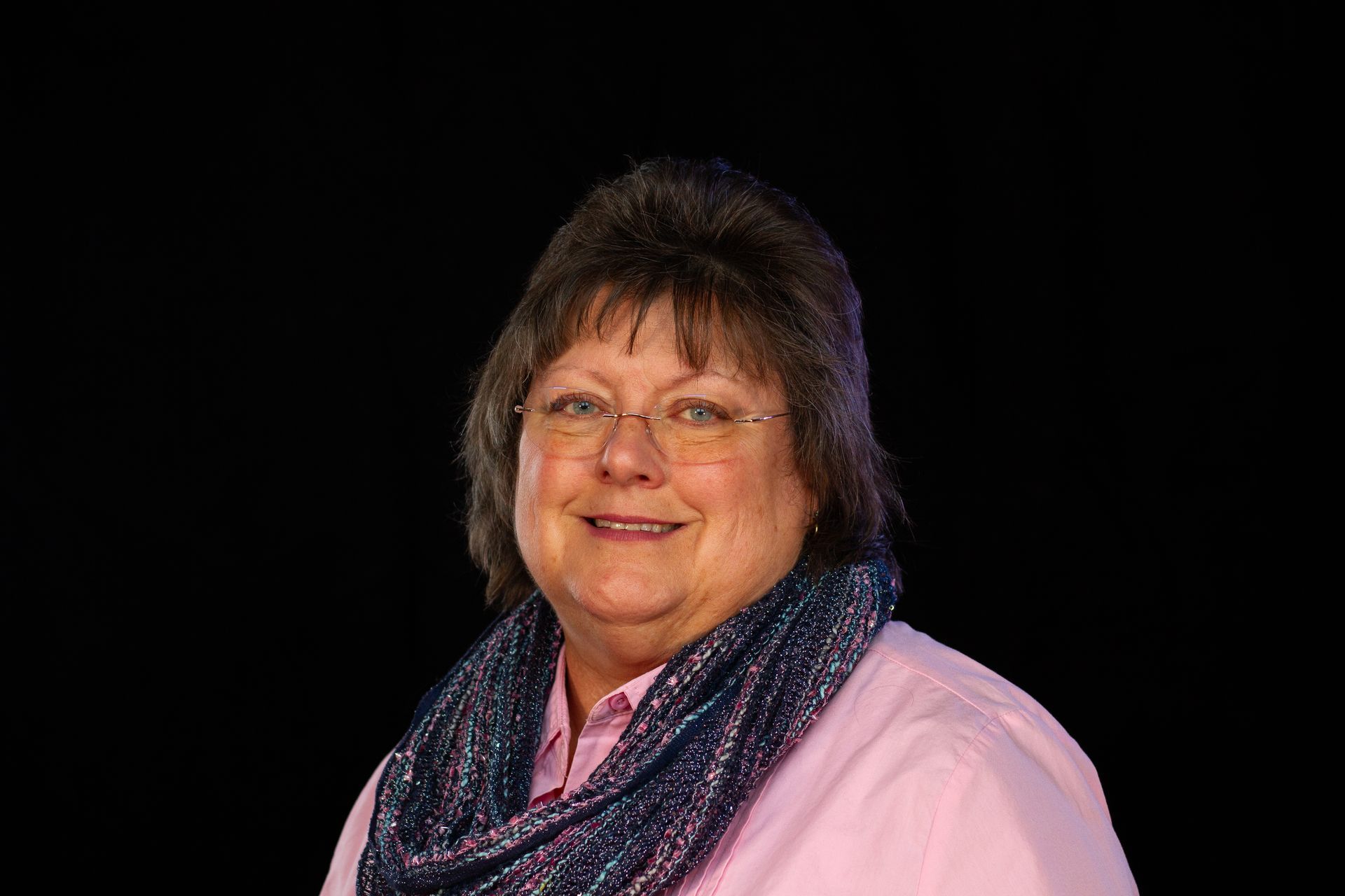 Woman with glasses smiling, wearing a pink shirt and blue scarf, against a black background.