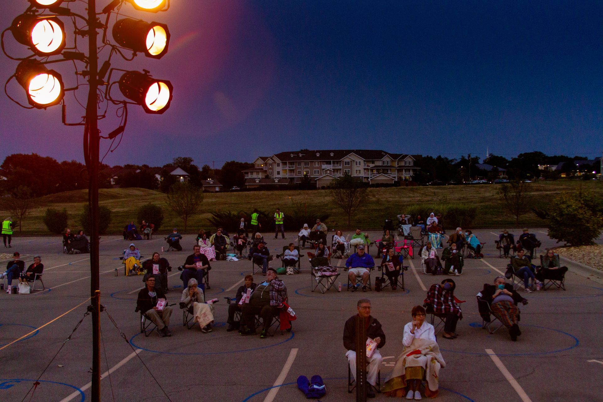 Parking lot set up for outdoor events during pandemic People in wheelchairs and chairs attend an outdoor event in a parking lot under lights at dusk.