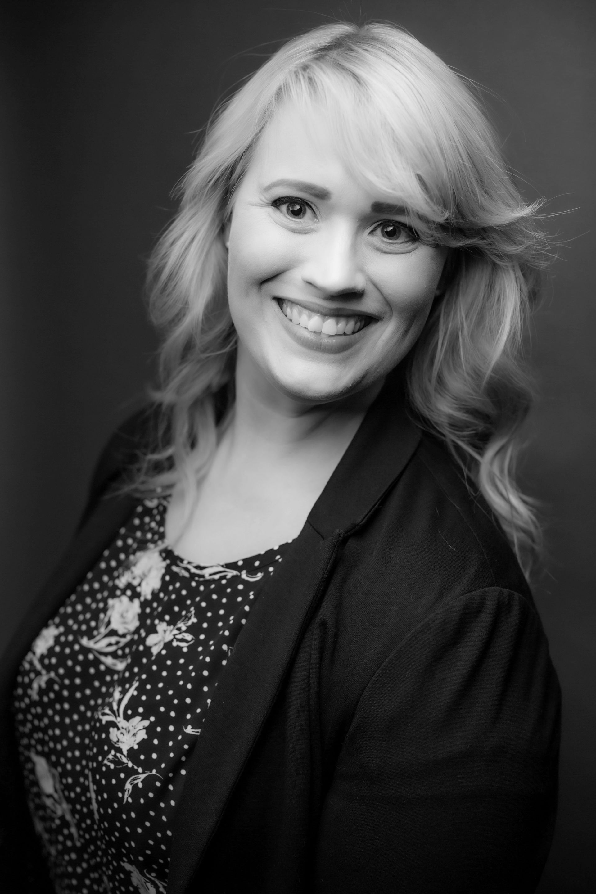 Woman with blonde hair smiles, wearing a blazer over a patterned top.