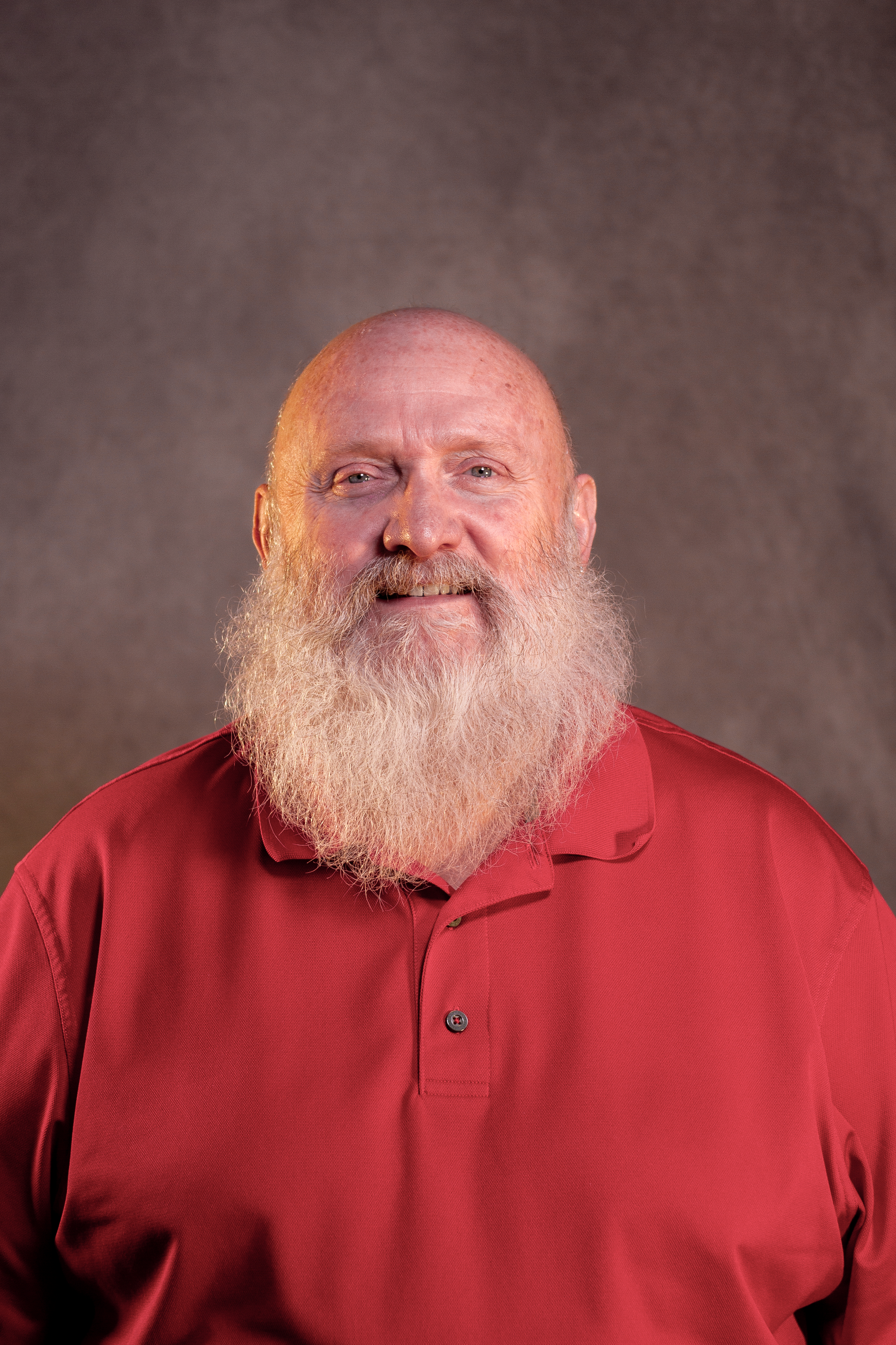 Bald man with a white beard smiles, wearing a red polo shirt, against a gray background.