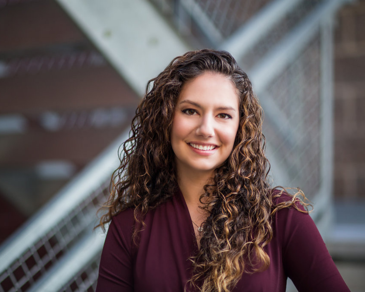 Smiling person with curly hair in a burgundy top, standing in front of a metal stairway.