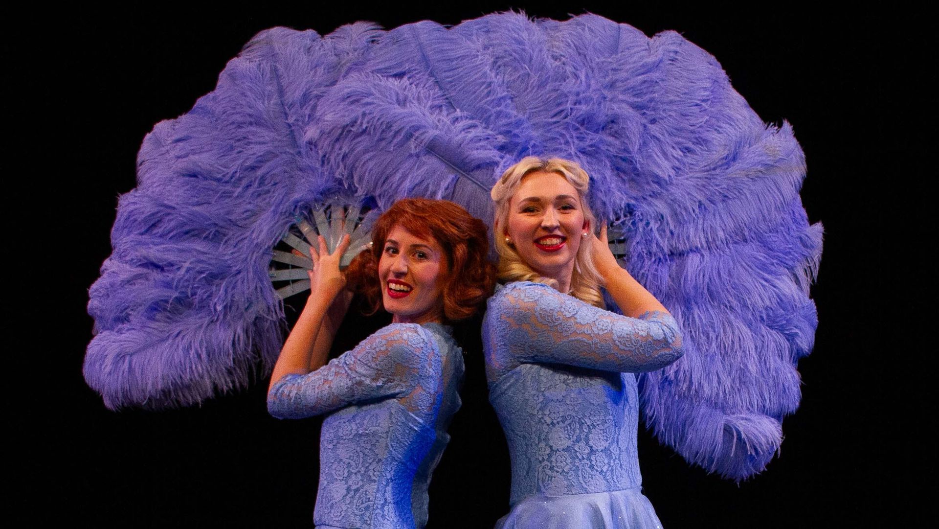 Two women in blue dresses, holding a large, blue feather fan, smiling, black background.