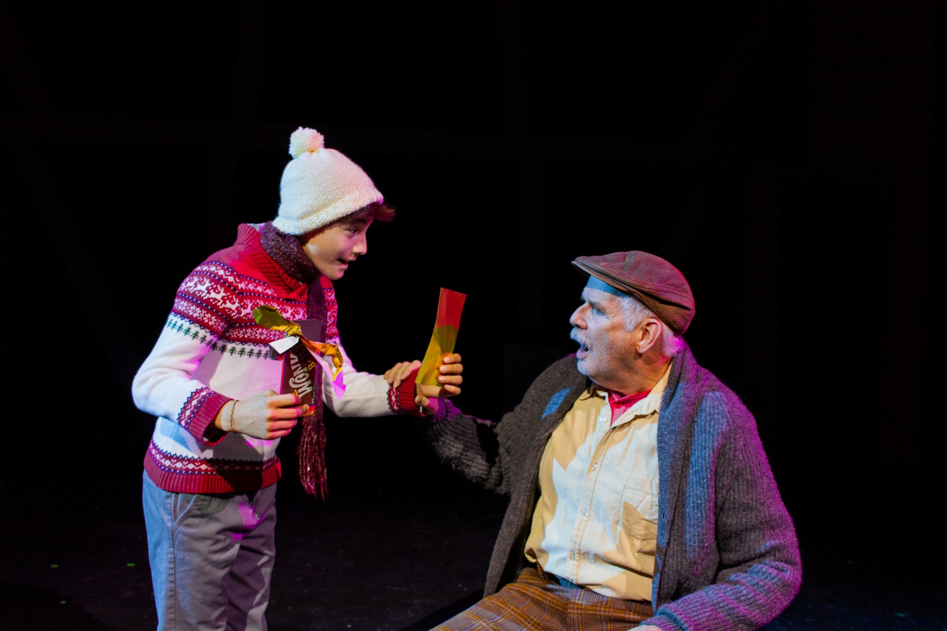 A person in a winter hat hands a colorful object to an older man. Stage setting with black background.