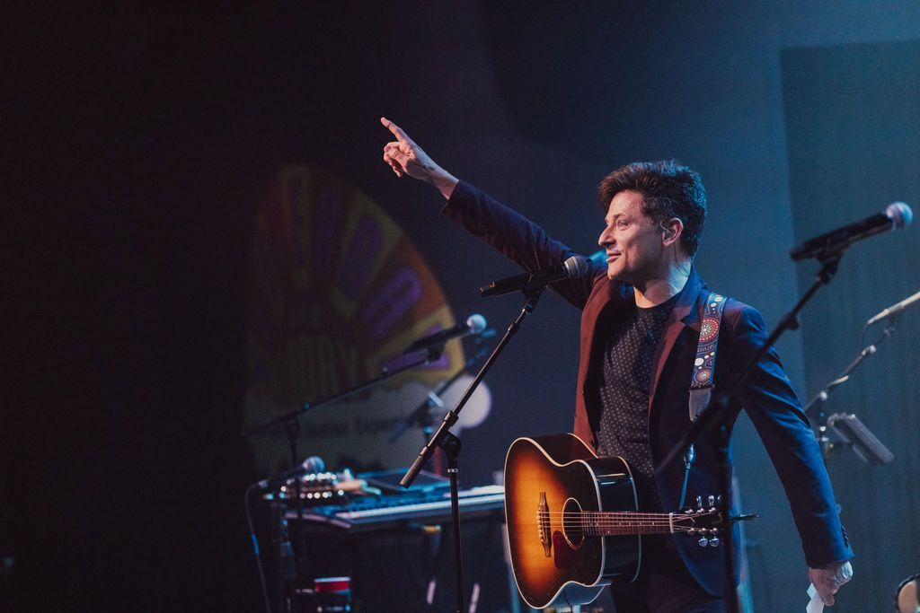 Man on stage with guitar, pointing up; stage lighting.