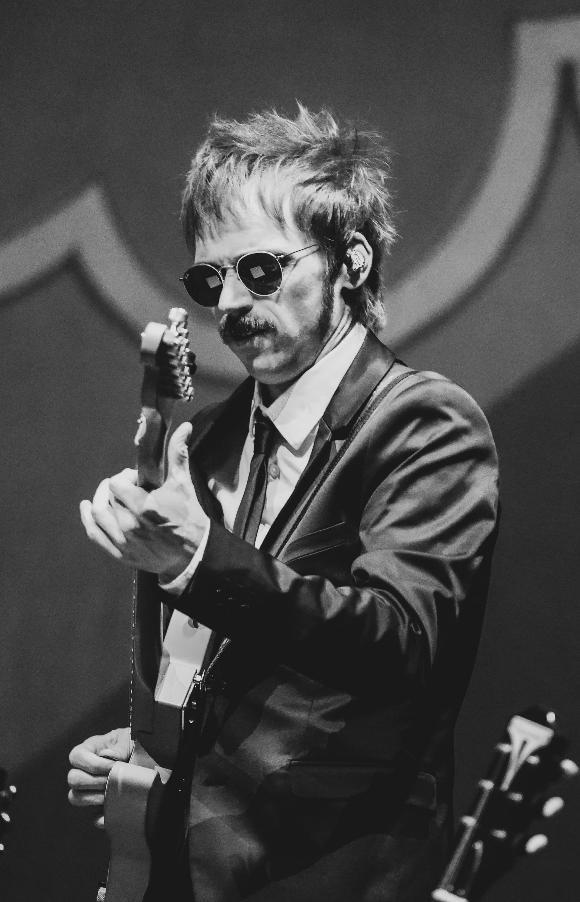 Musician playing guitar, wearing sunglasses and a suit, on stage.