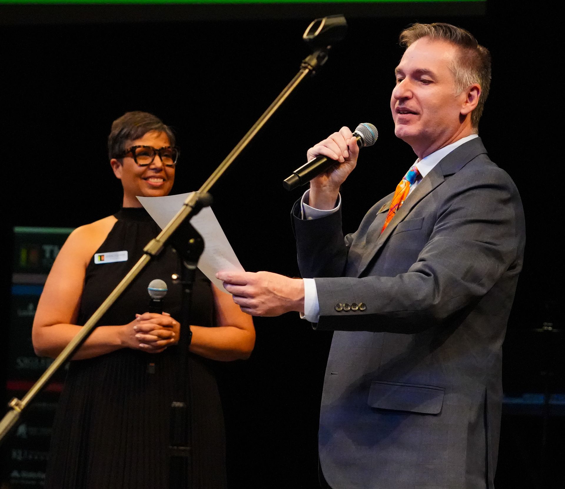 Man in a suit speaks into a microphone, holding papers. A woman stands beside him, smiling, holding a microphone.